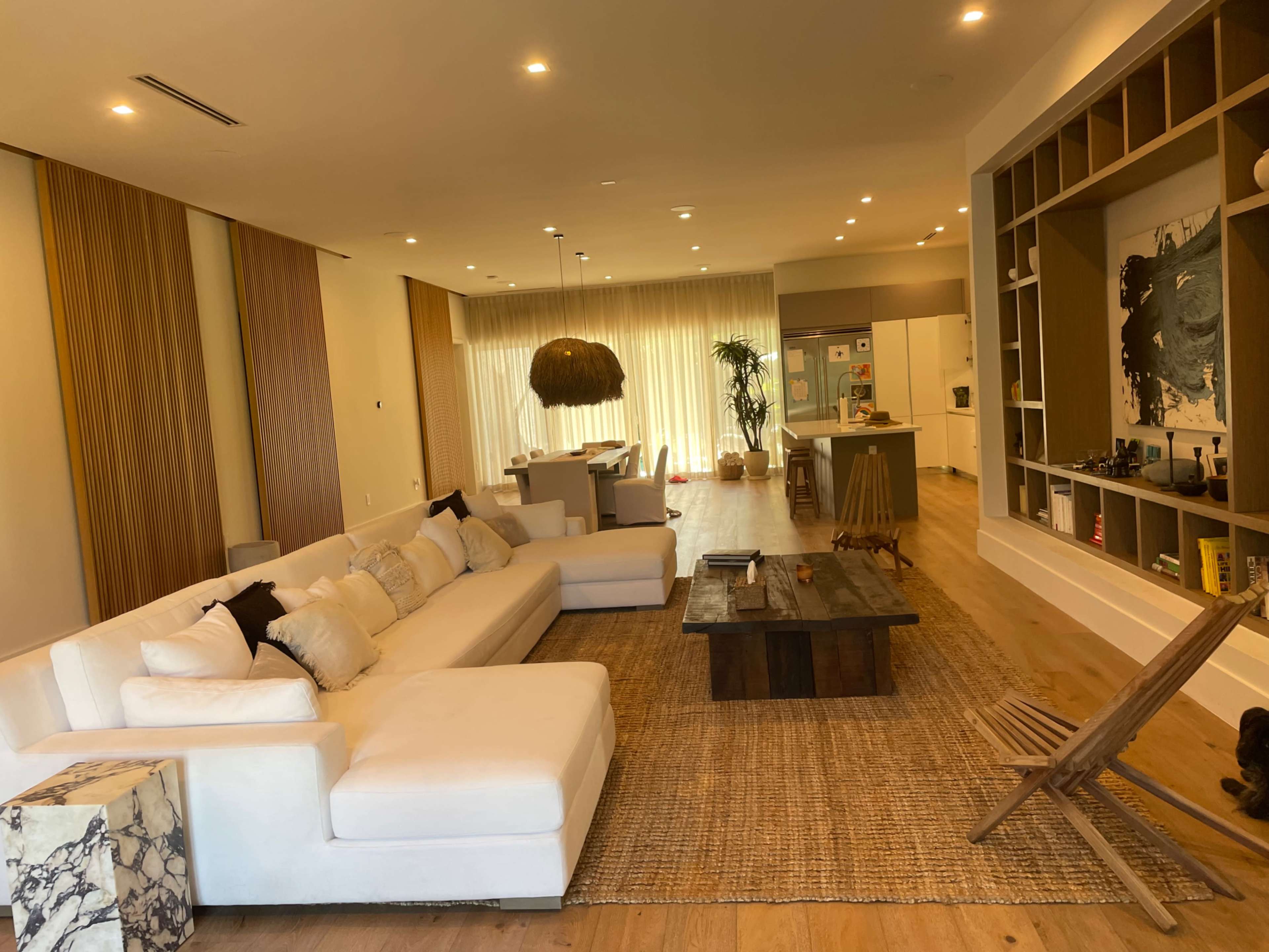 The image shows a modern living room with a large white sectional sofa, a wooden coffee table, and a woven rug, leading into a kitchen area with multiple plants and ample natural light.