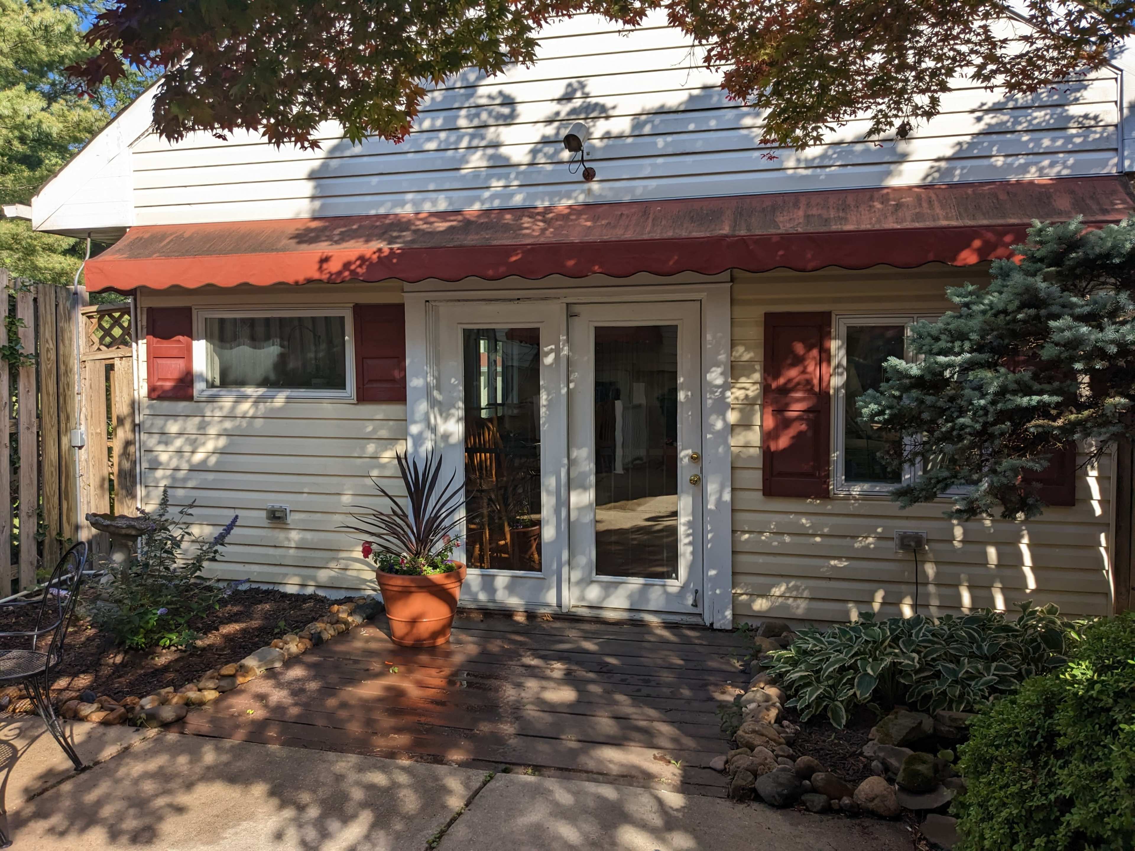 A small outdoor patio area with a white house, a red awning over a glass door, and landscaped borders featuring plants and stones.