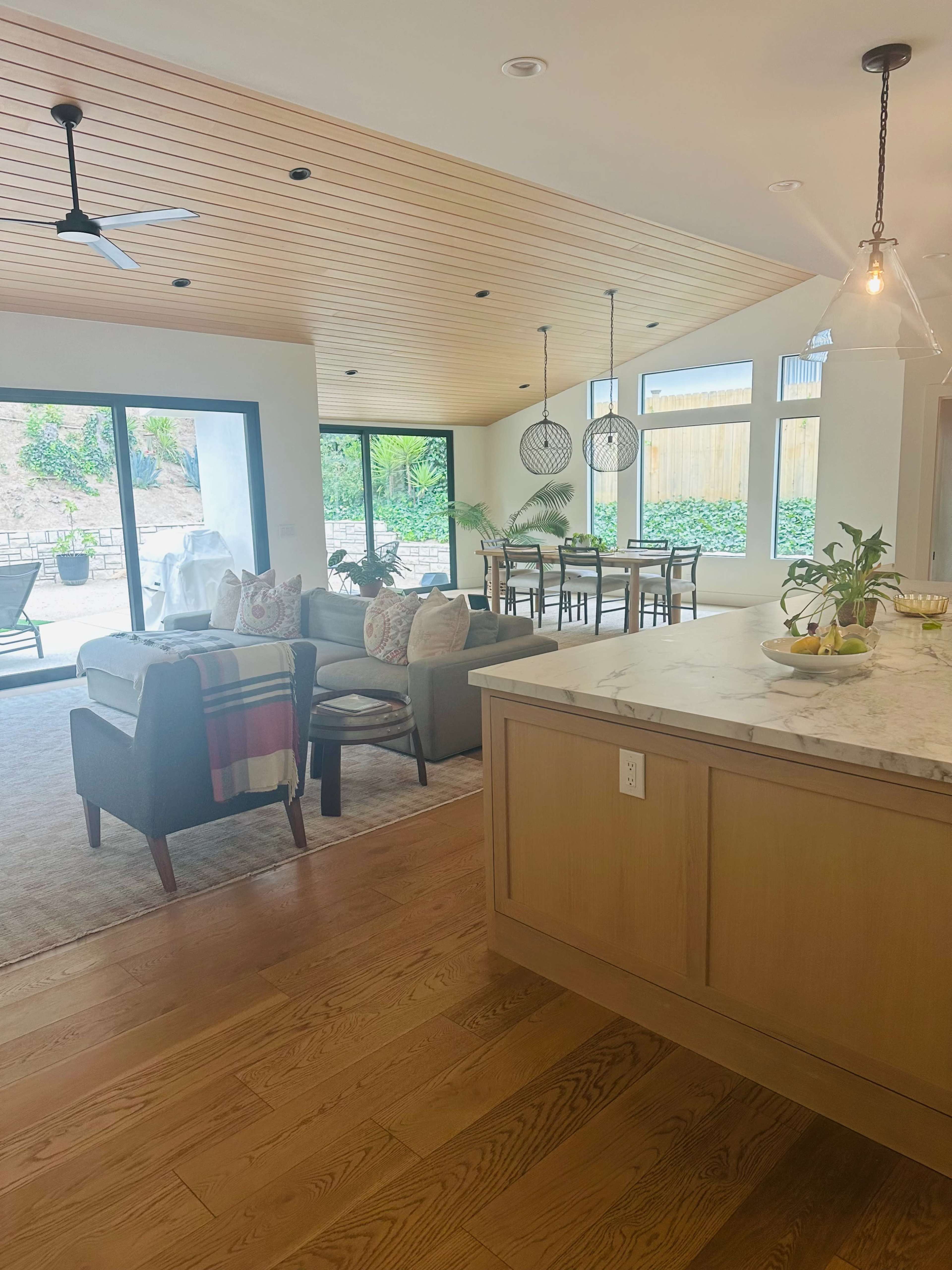 The image shows a bright and modern living and dining area with a kitchen island, featuring wood ceilings, large windows, and a view of an outdoor space.