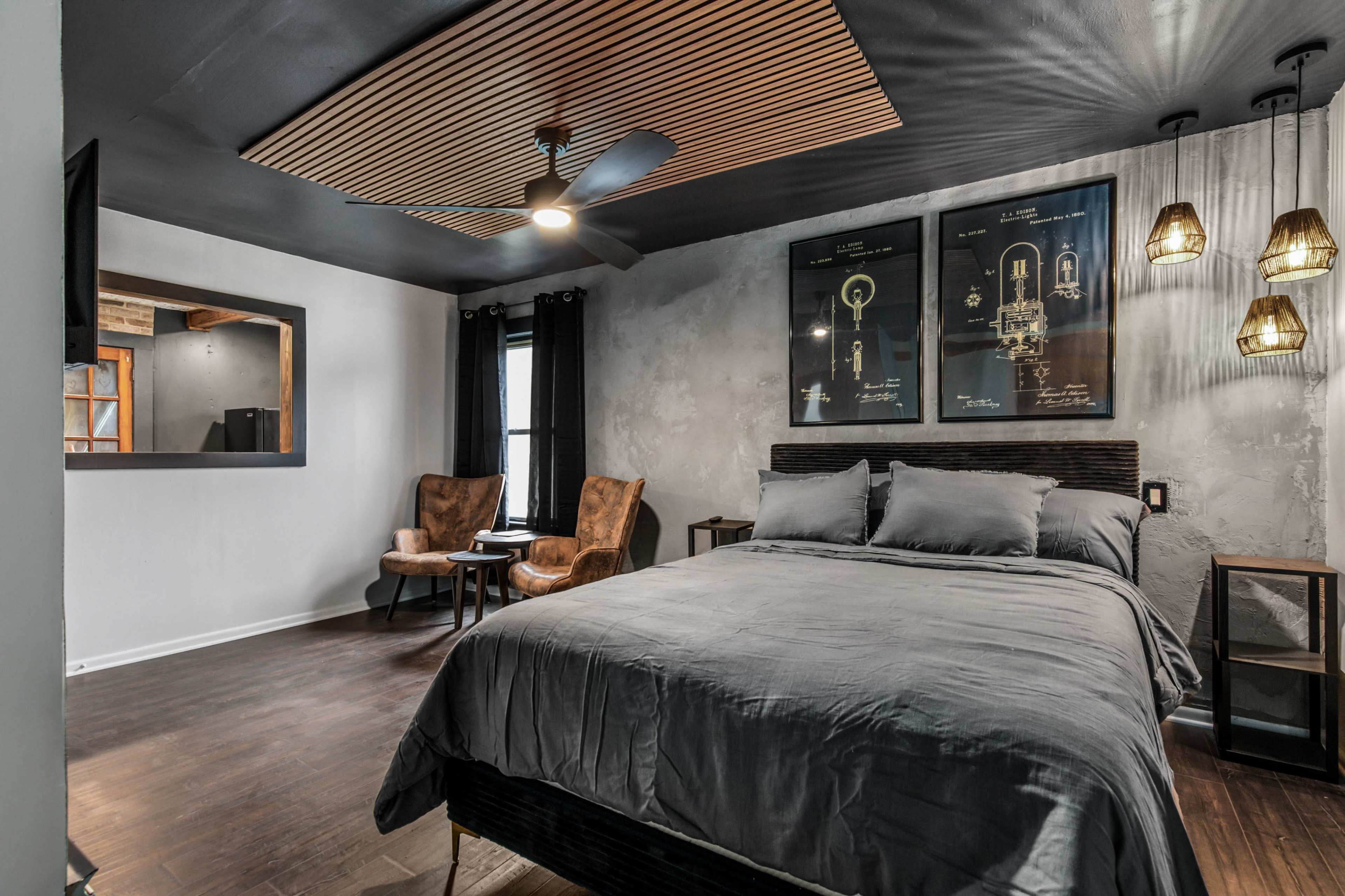 A modern bedroom featuring a large bed with gray bedding, two chairs, and a decorative wall with framed art and pendant lights.
