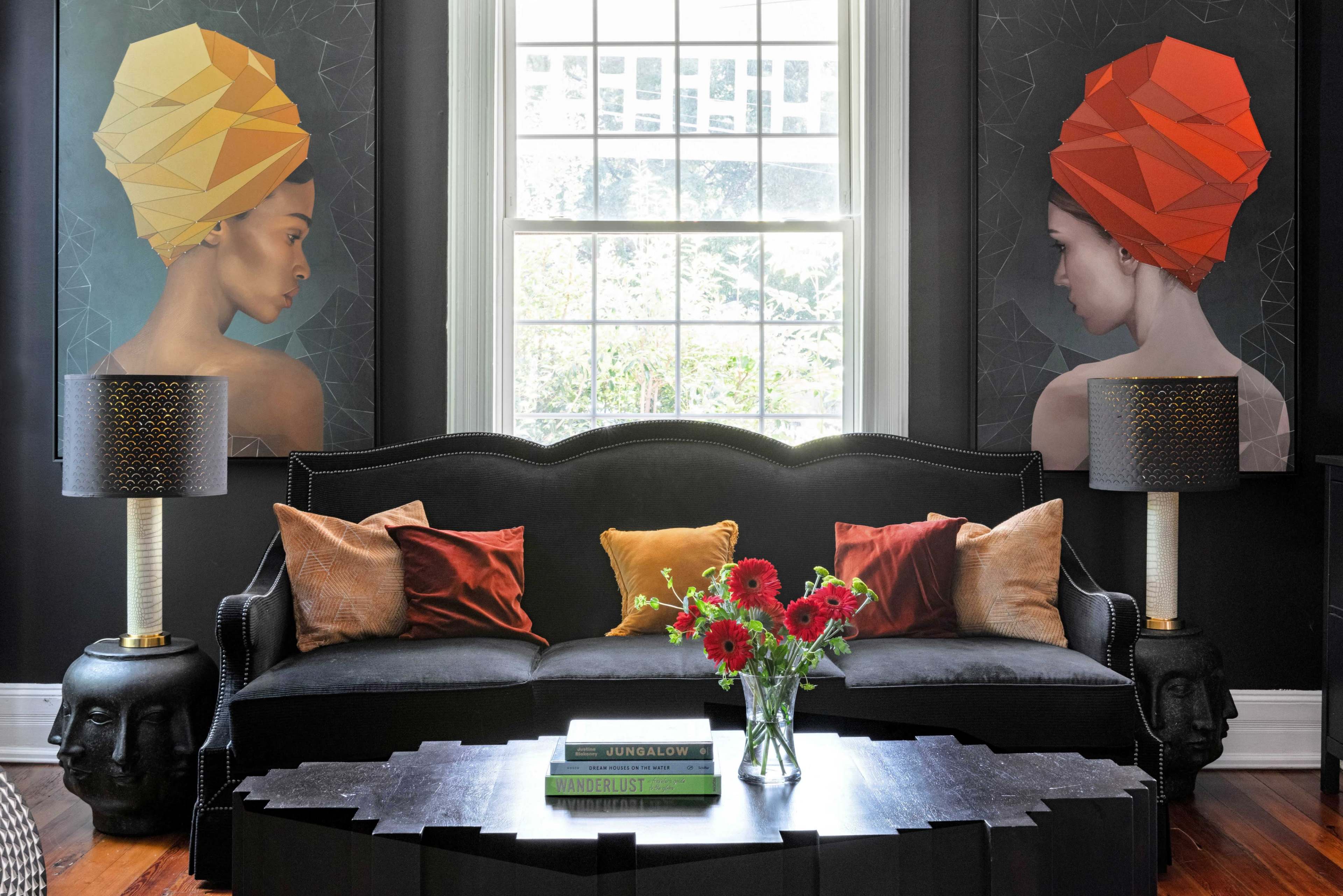 A modern living room features a black velvet sofa adorned with colorful cushions, two artistic wall portraits, and a coffee table with a vase of flowers.