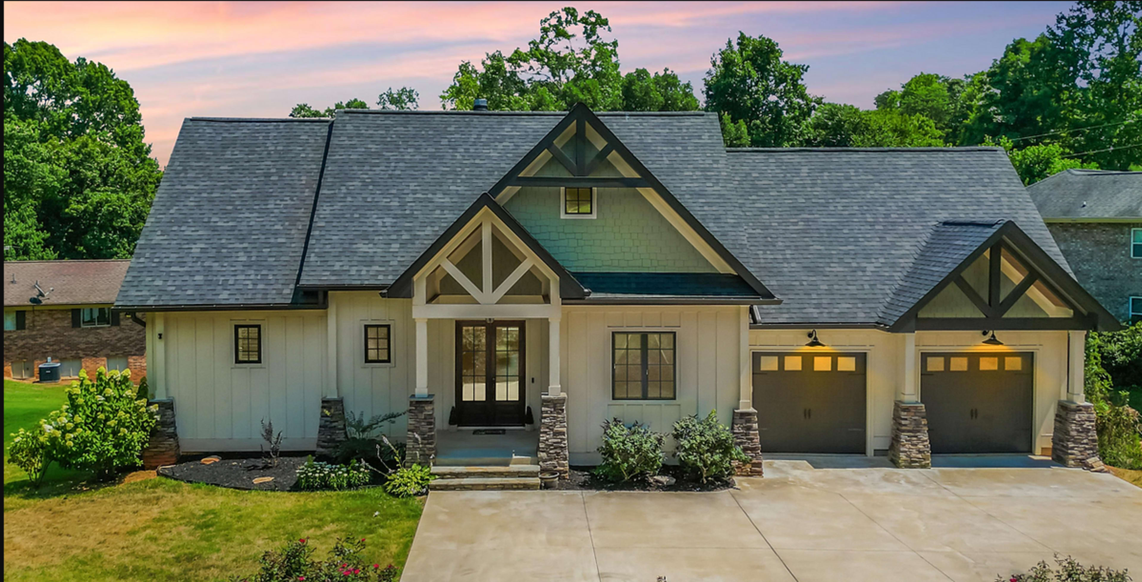 The image shows a modern, single-story house with a steeply pitched roof, two garage doors, and a landscaped front yard.