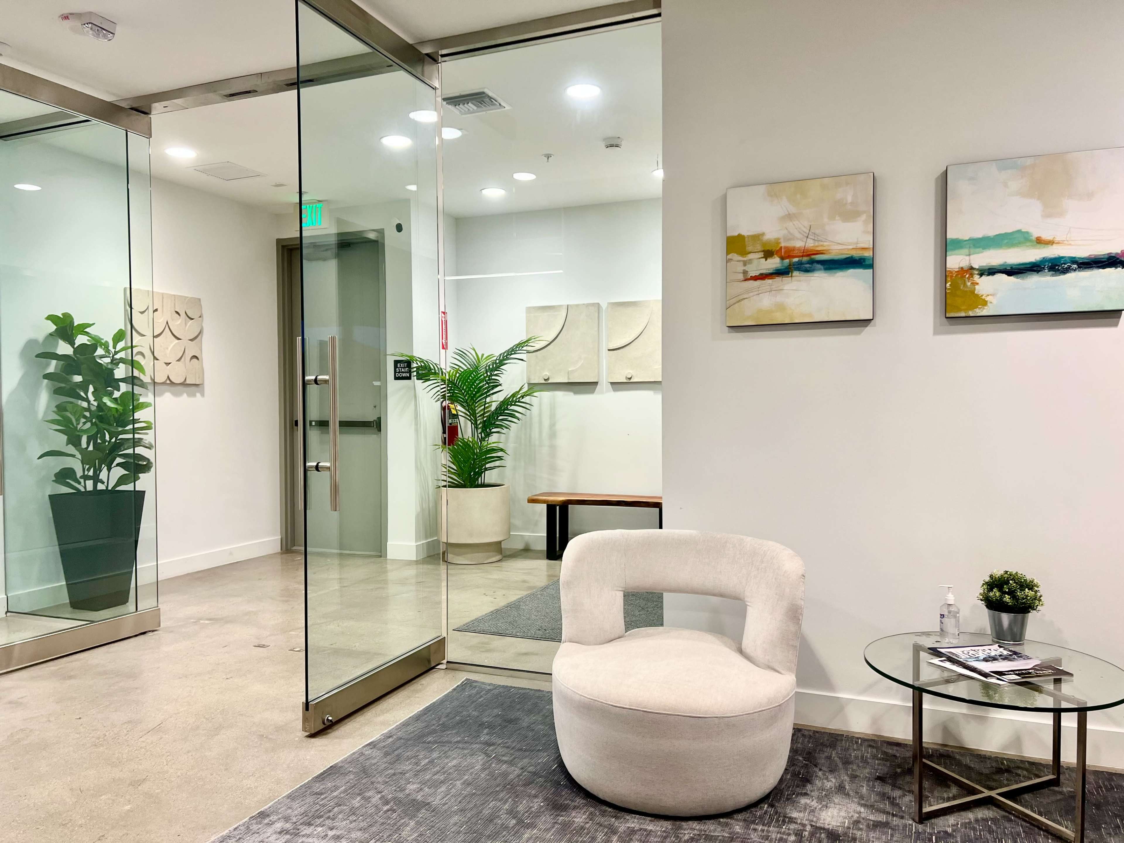 A modern indoor waiting area with a round white chair, a small table, and a potted plant, featuring artwork on the walls and glass doors leading to another room.