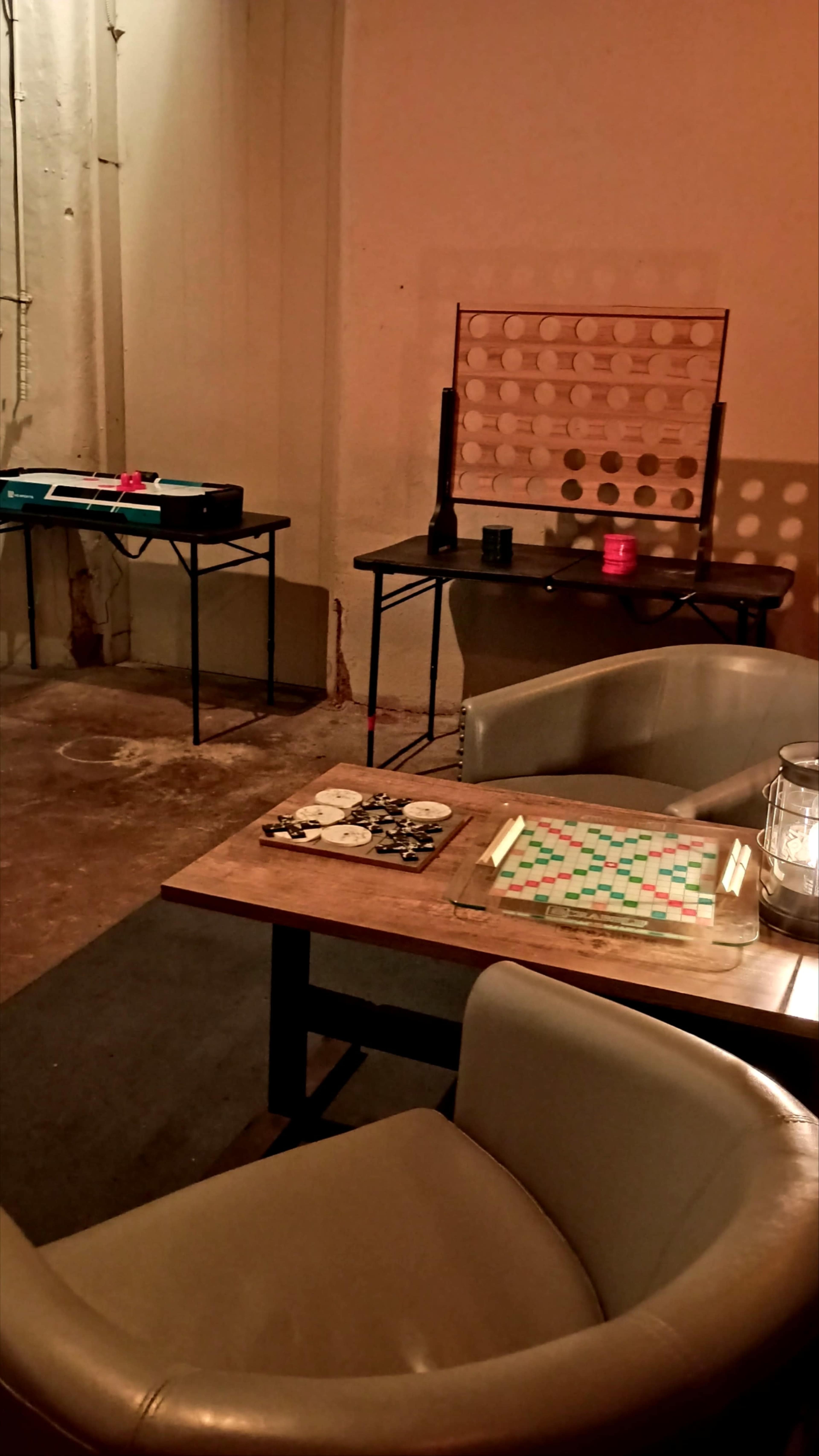 The image shows a dimly lit room with two tables: one set up with a Scrabble game and the other with a board for Connect Four, alongside a couple of chairs and a lighted lantern.