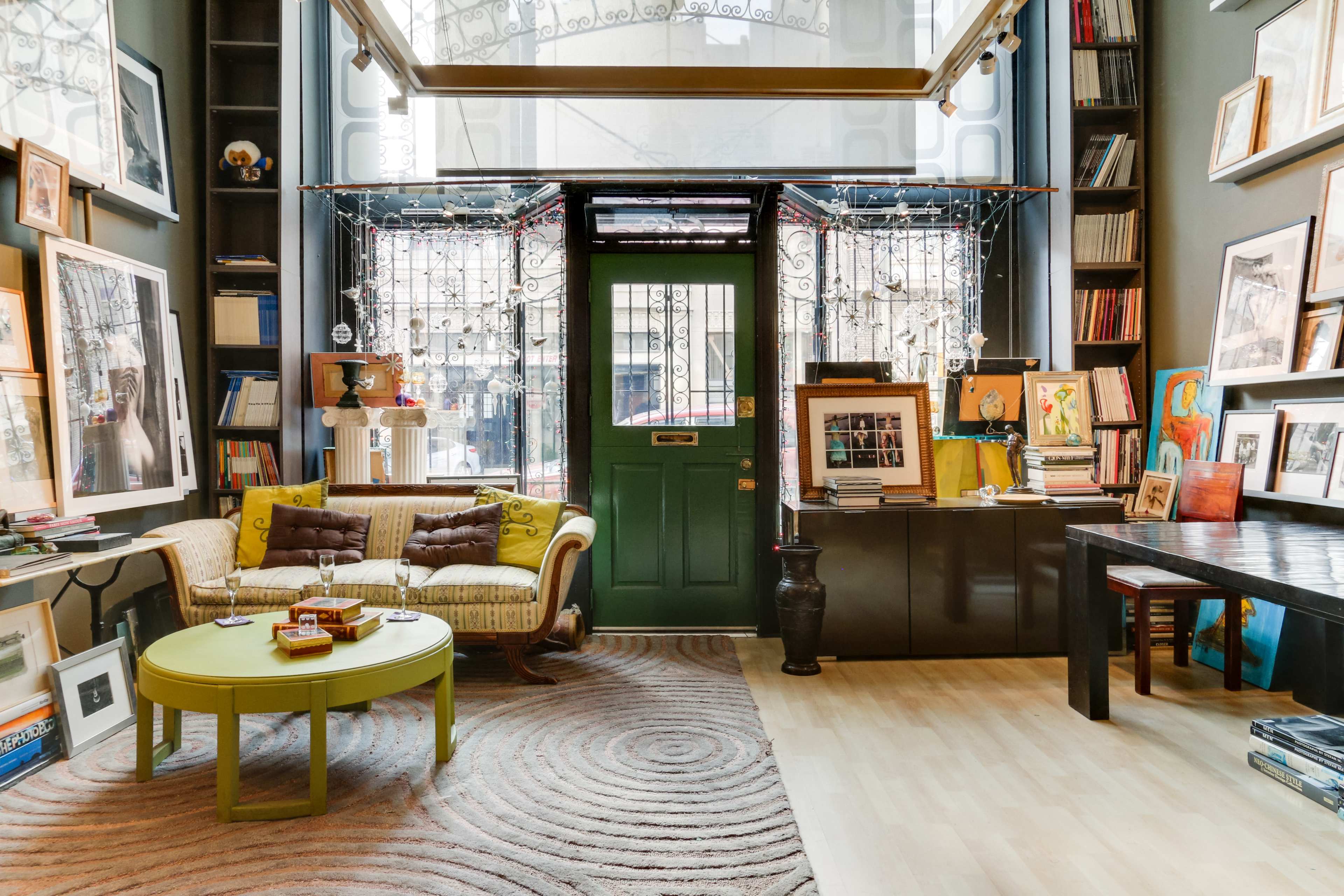 A cozy interior of a room featuring a green door, a yellowish sofa with a circular coffee table, and walls adorned with framed pictures and shelves filled with books and art.
