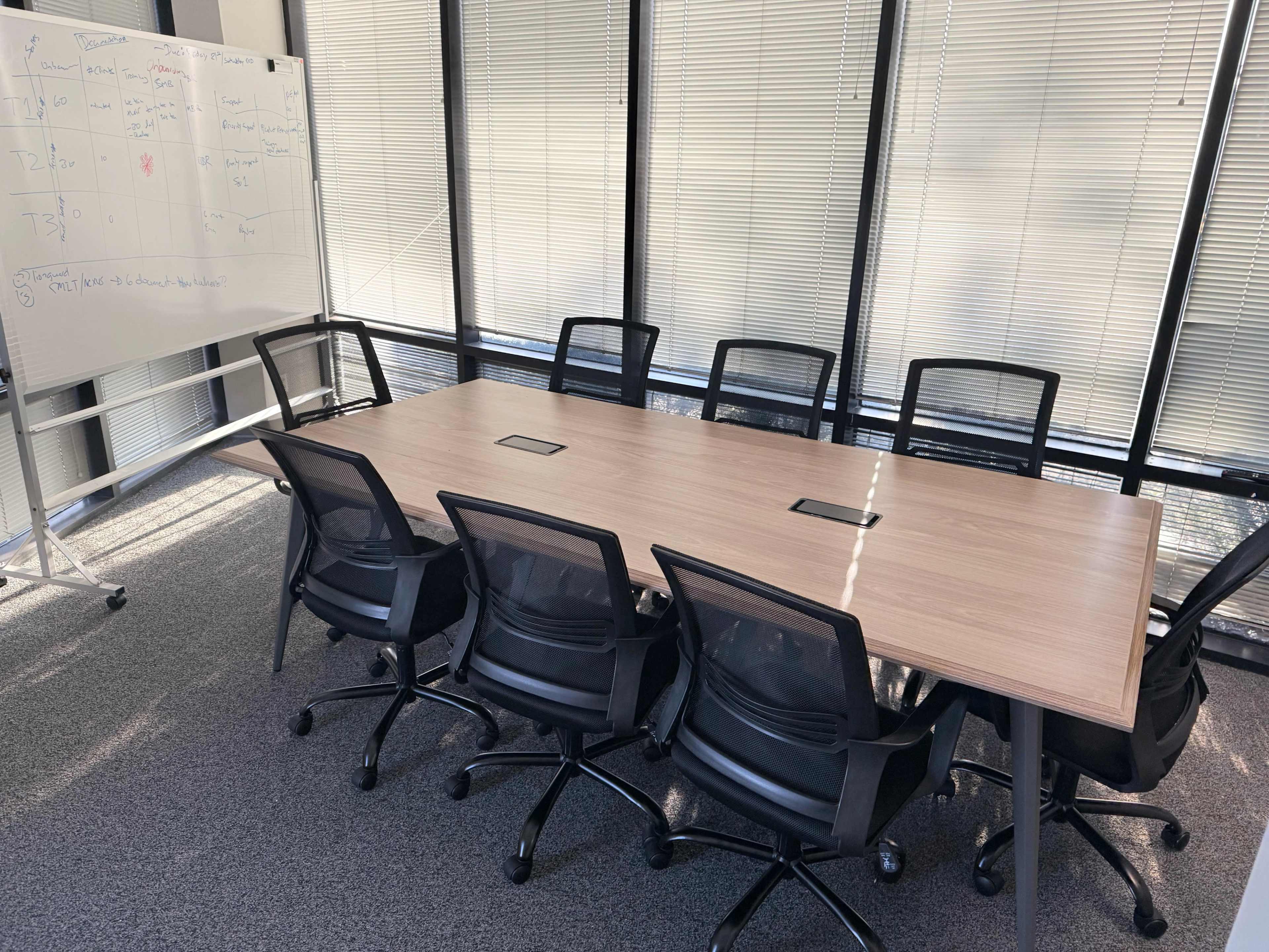 A modern conference room features a long wooden table surrounded by black ergonomic chairs and a whiteboard in the background.