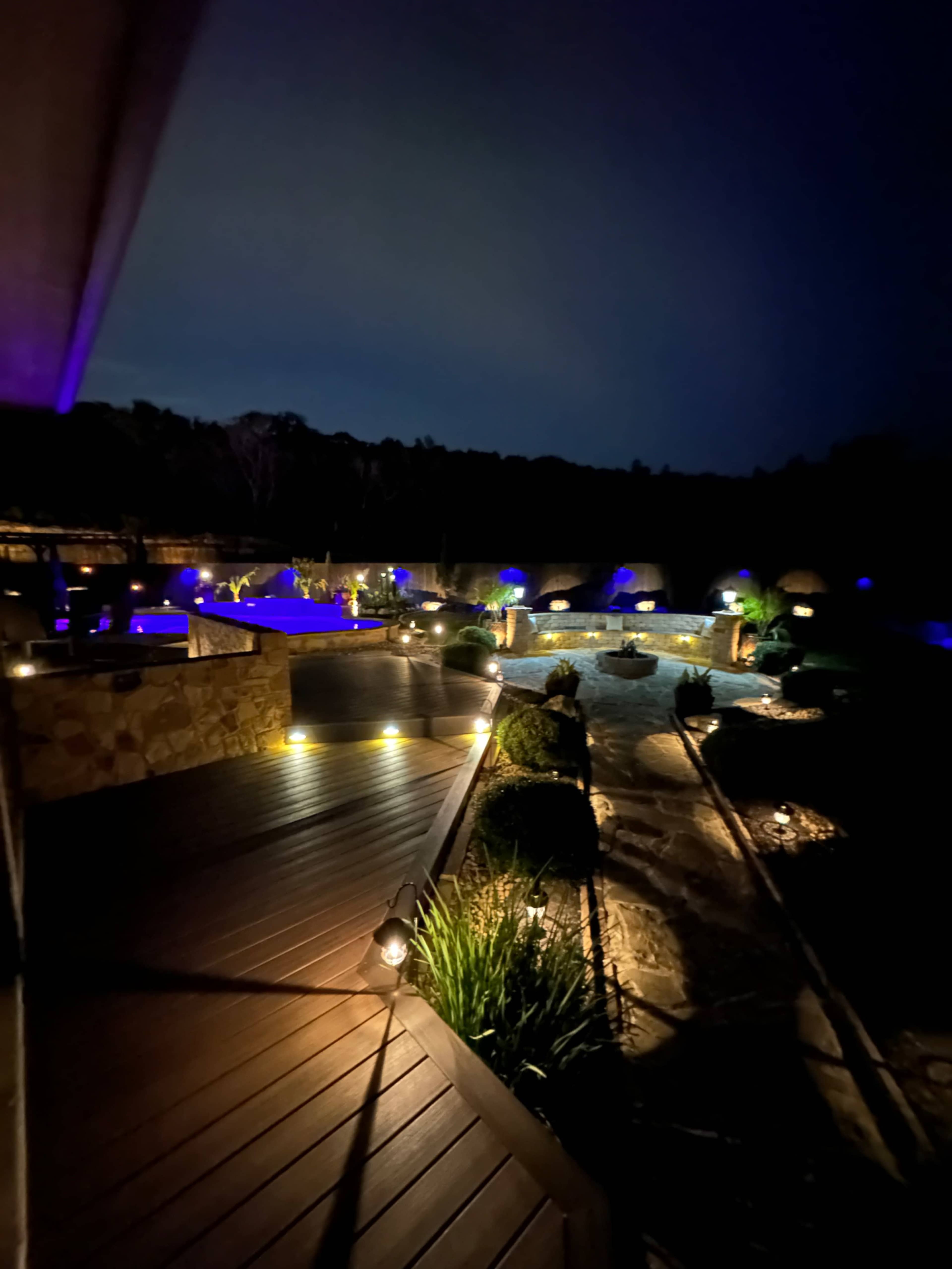The scene features a well-lit outdoor area at night, showcasing landscaped gardens with pathways, water features, and ambient lighting.