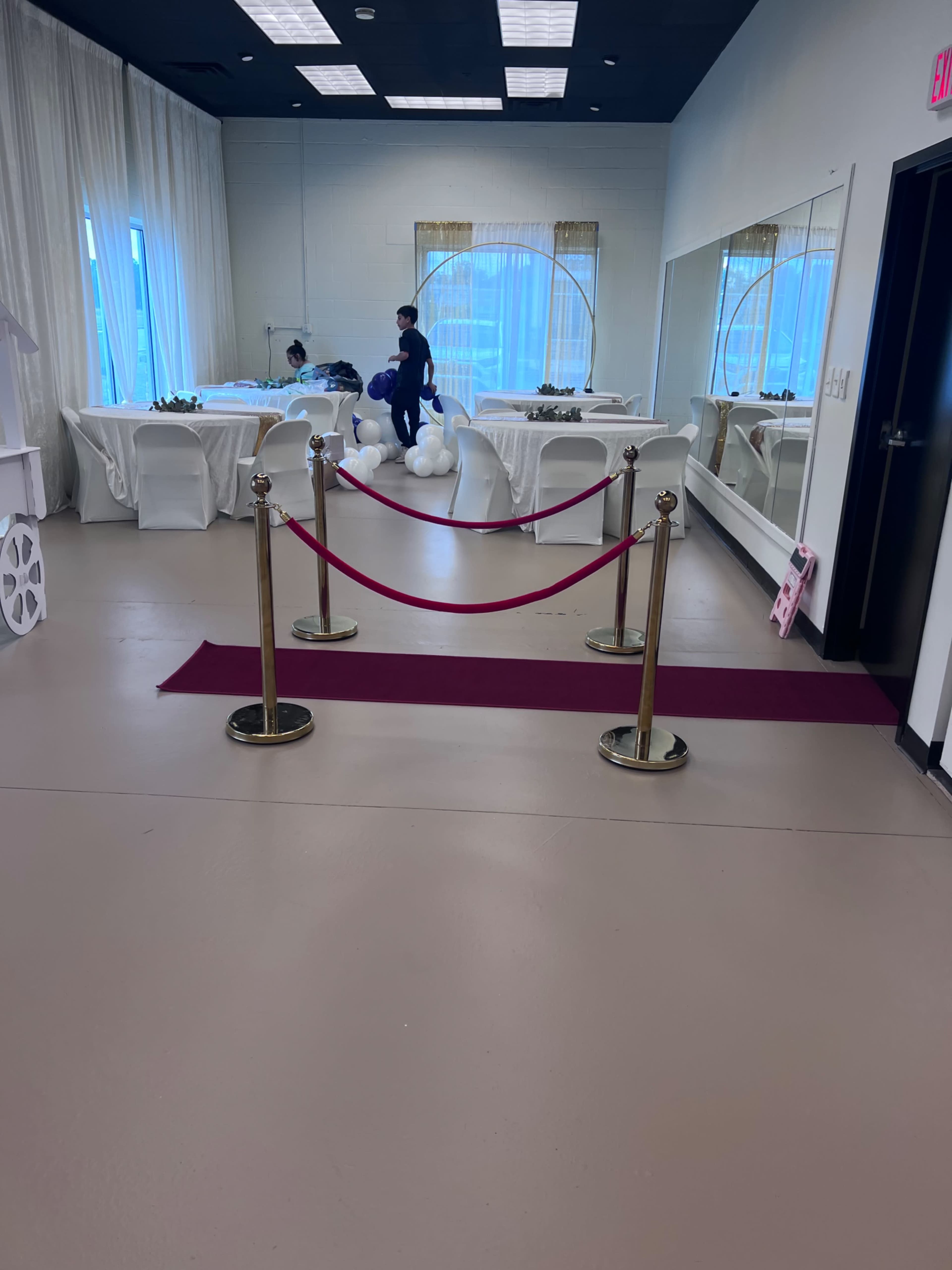 An indoor event space with white tables and chairs, a red carpet leading to a sectioned area, and a person arranging decorations nearby.