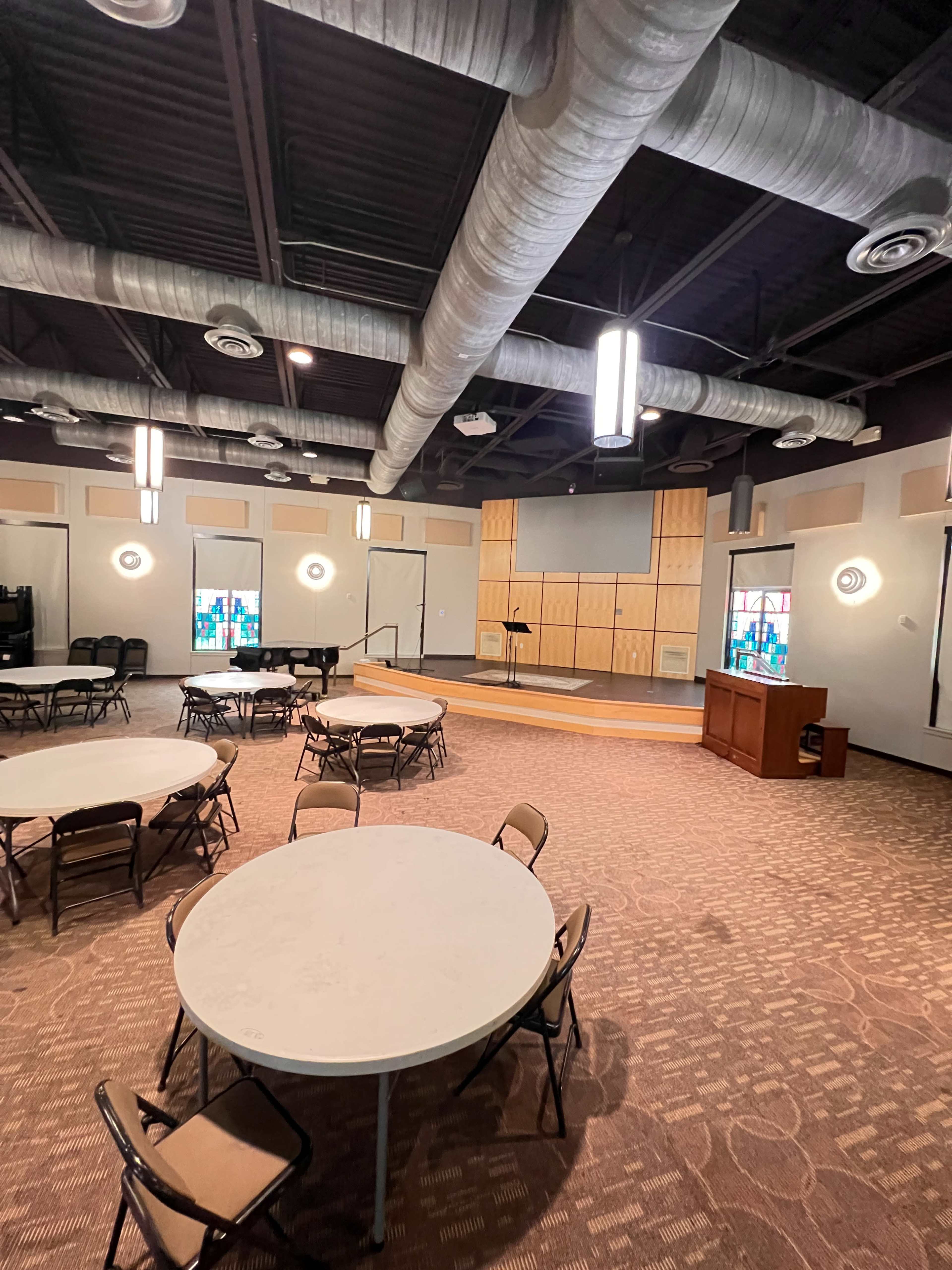 The image shows a large, empty room set up for an event, featuring multiple round tables and a stage at the front with a wooden podium.