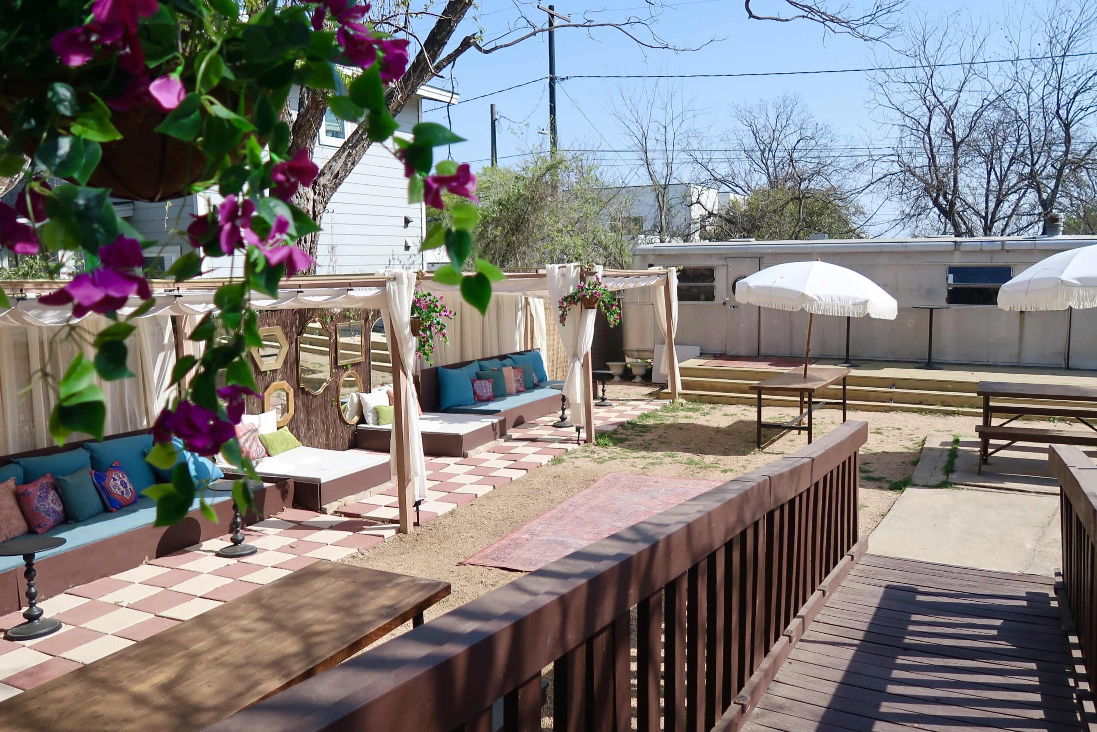The image shows a garden patio featuring flower-adorned seating areas, a shaded table, and a white umbrella, surrounded by a brown wooden railing.