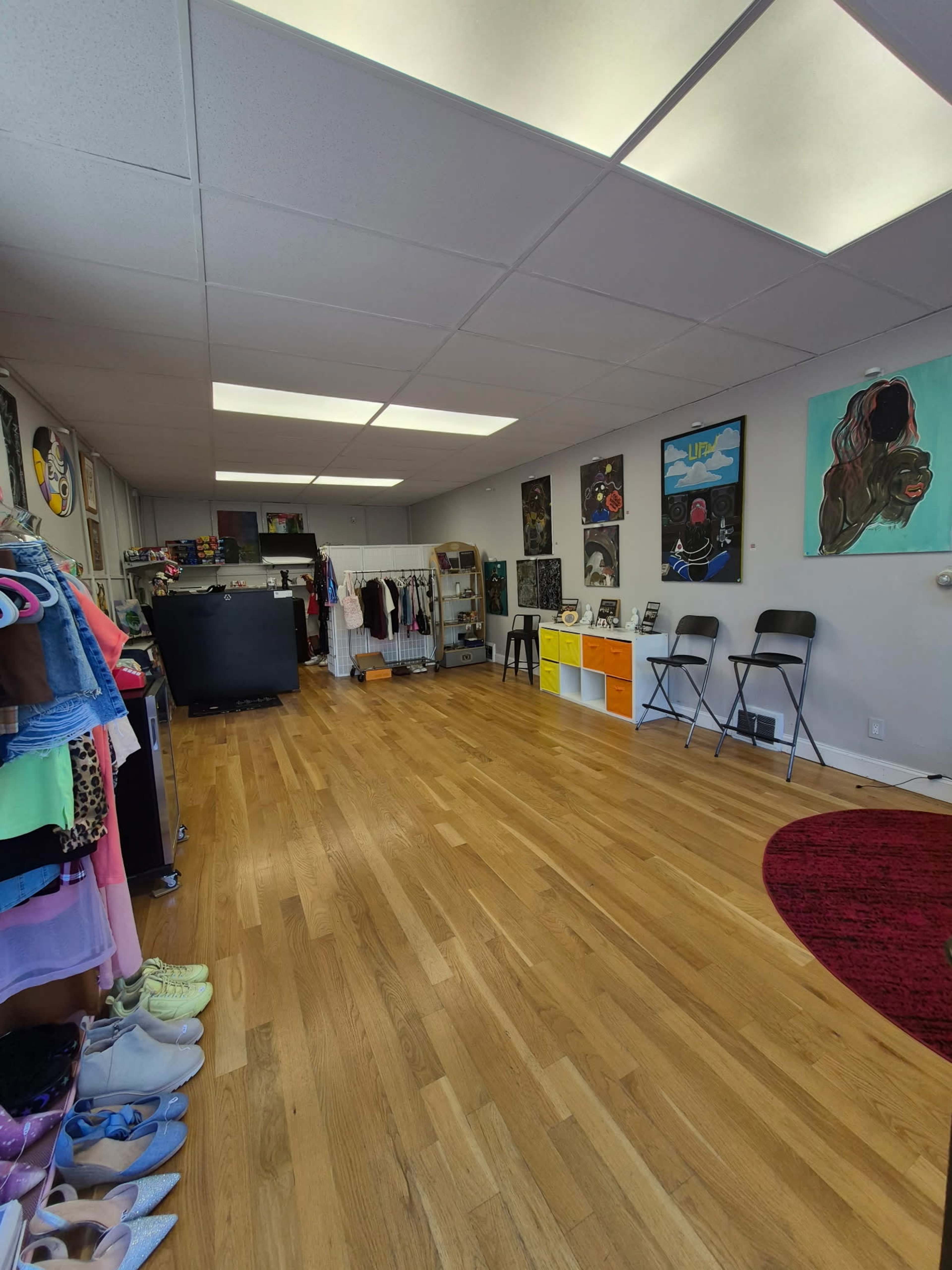 The image shows a retail space with wooden flooring, clothing racks, chairs, and colorful artwork on the walls.