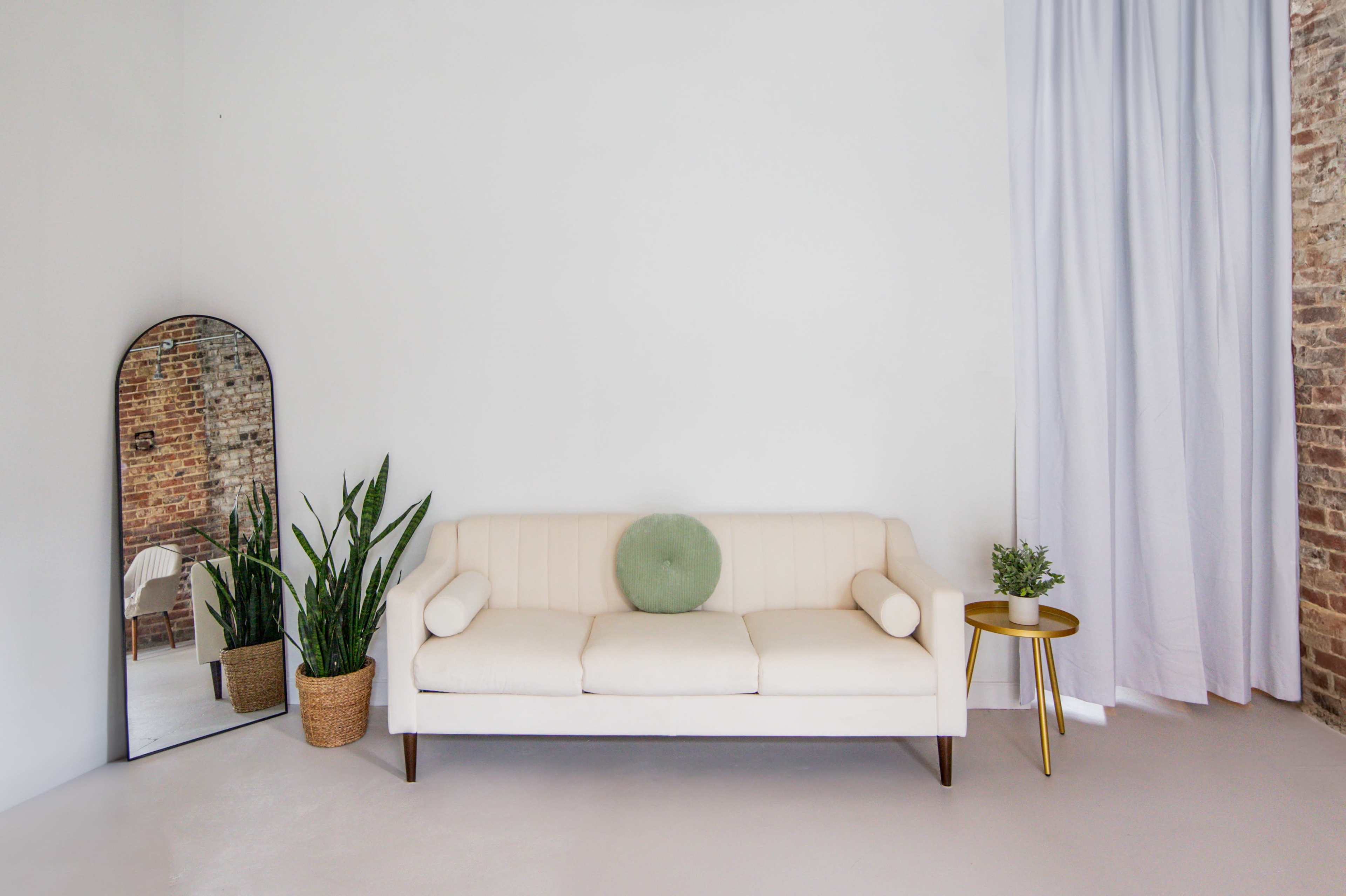 A light-colored sofa is placed against a plain wall, accompanied by a tall plant, a round mirror, and a small side table with a potted plant.