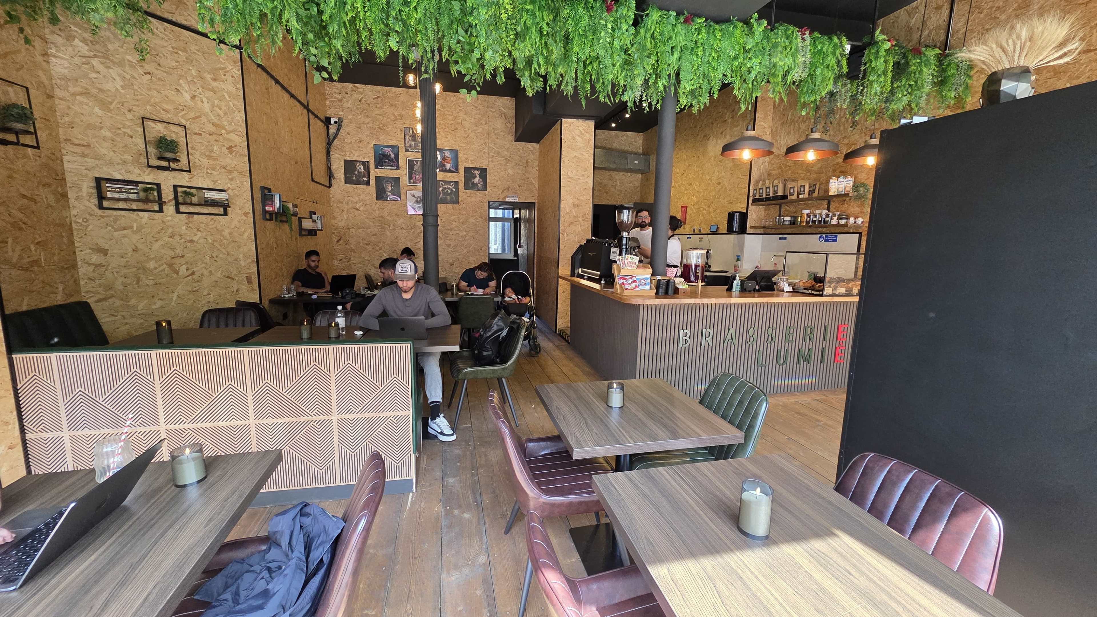 The image shows a modern café interior with wooden tables, green plants hanging from the ceiling, and customers sitting at various tables, some working on laptops.