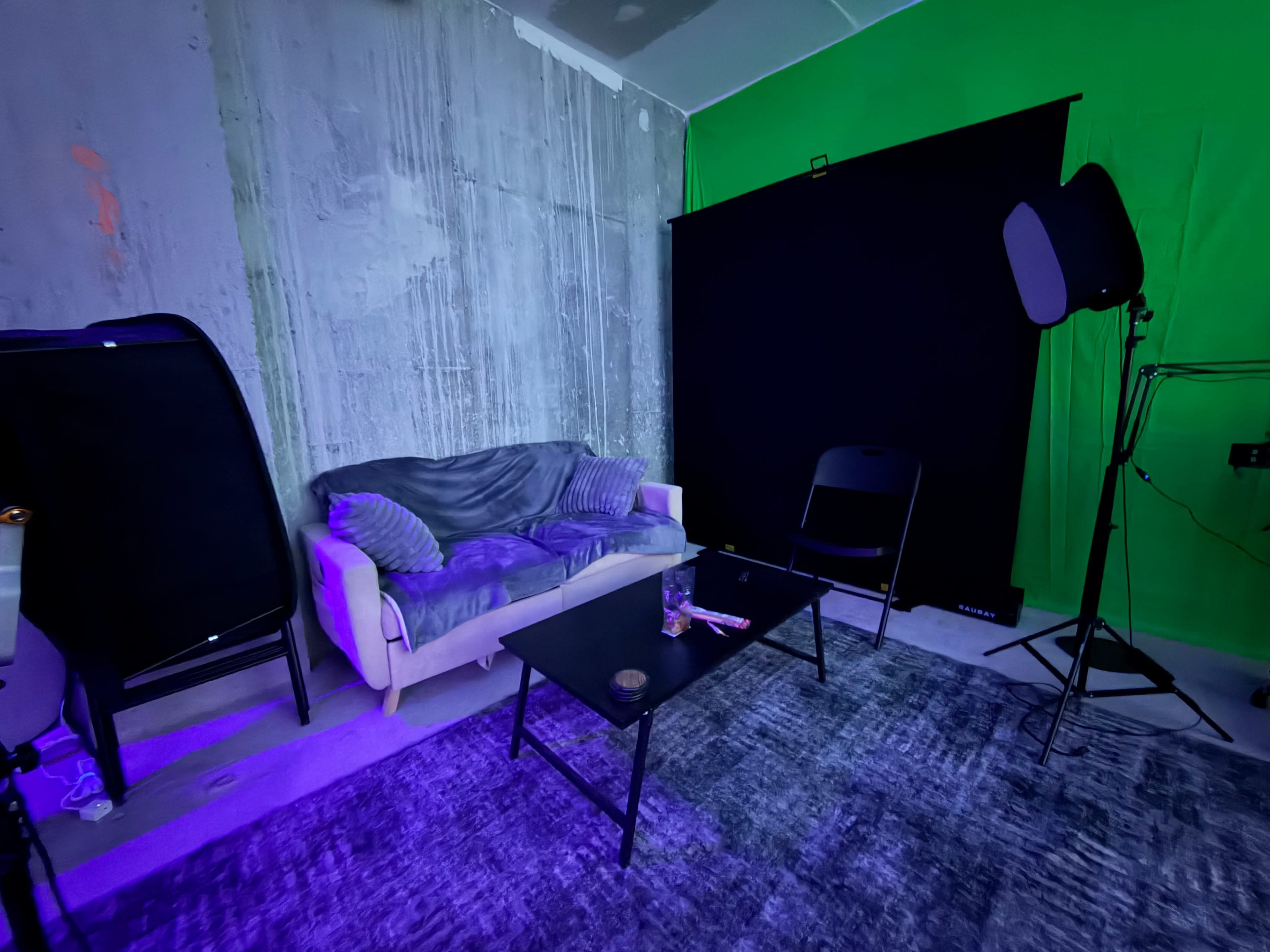 The image shows a small studio space with a gray sectional sofa, a black chair, and a black backdrop, all under dim purple lighting.