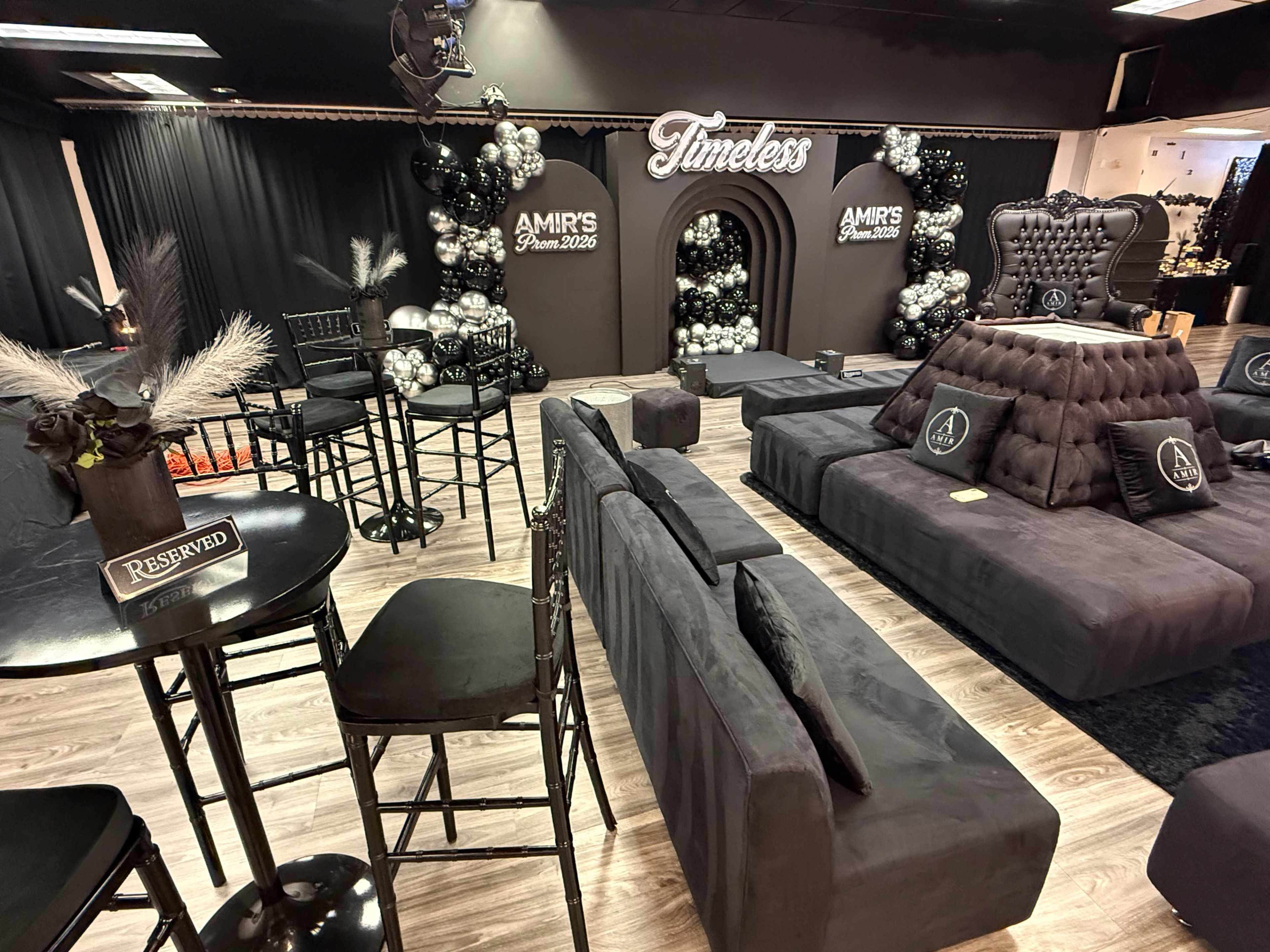 The image shows a stylish event space decorated in black and silver with plush seating, a backdrop featuring the word "Timeless," and decorative balloons.