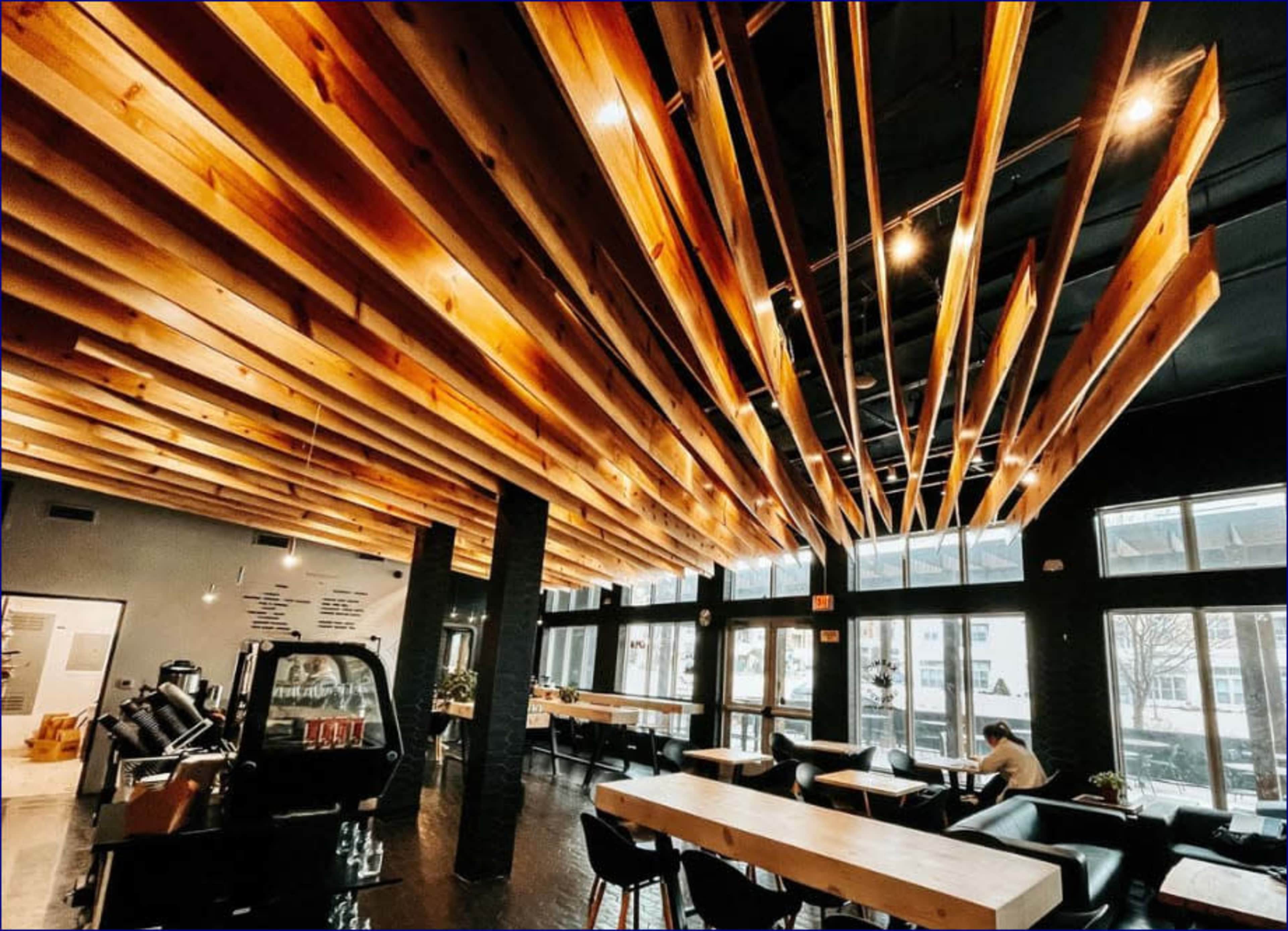 The interior of a modern café features a ceiling with wooden beams and pendant lights, large windows, and wooden tables.