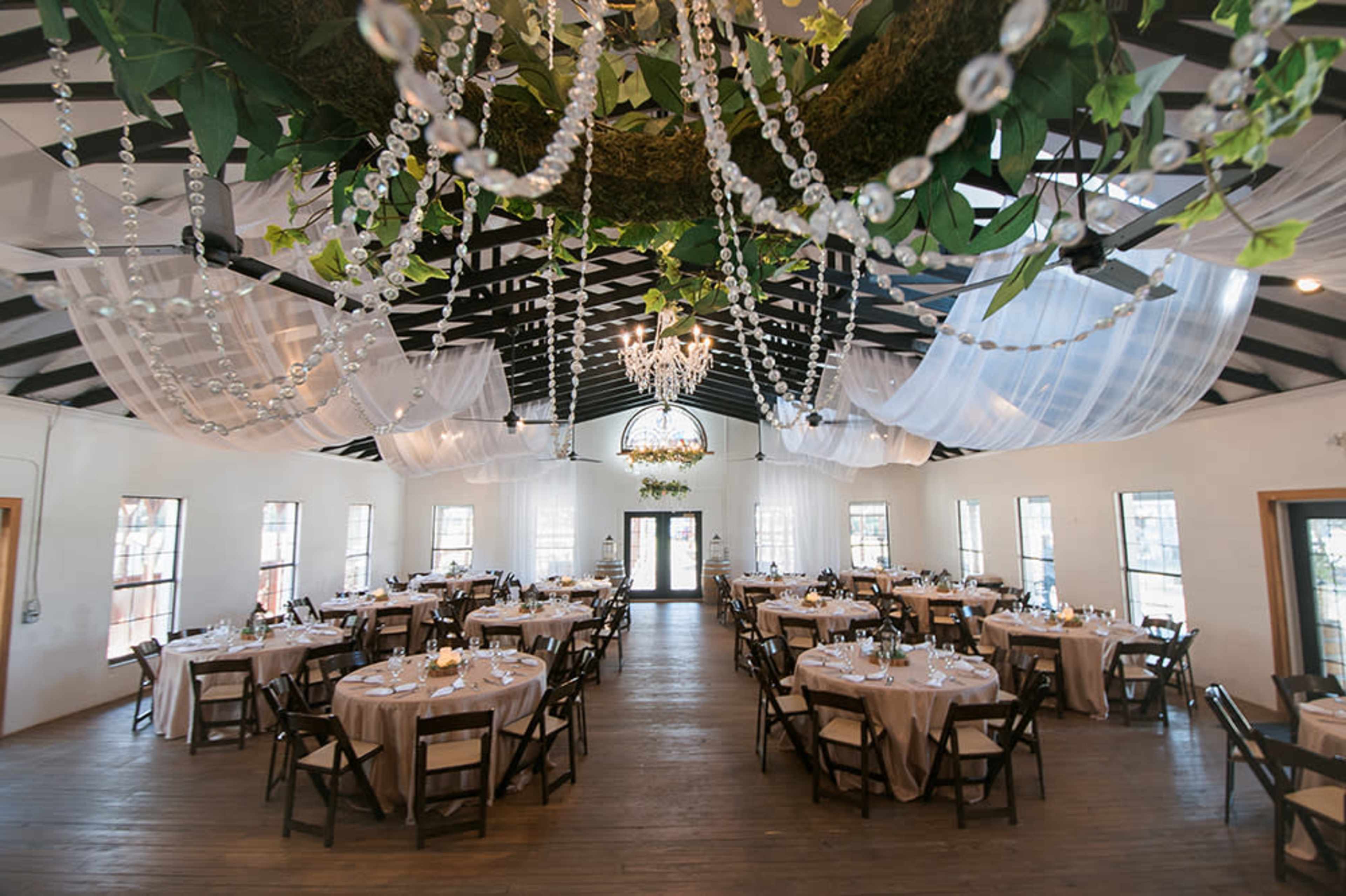 The image shows an elegantly decorated event space with tables set for dining, surrounded by draped fabric and a chandelier made of greenery and crystals.
