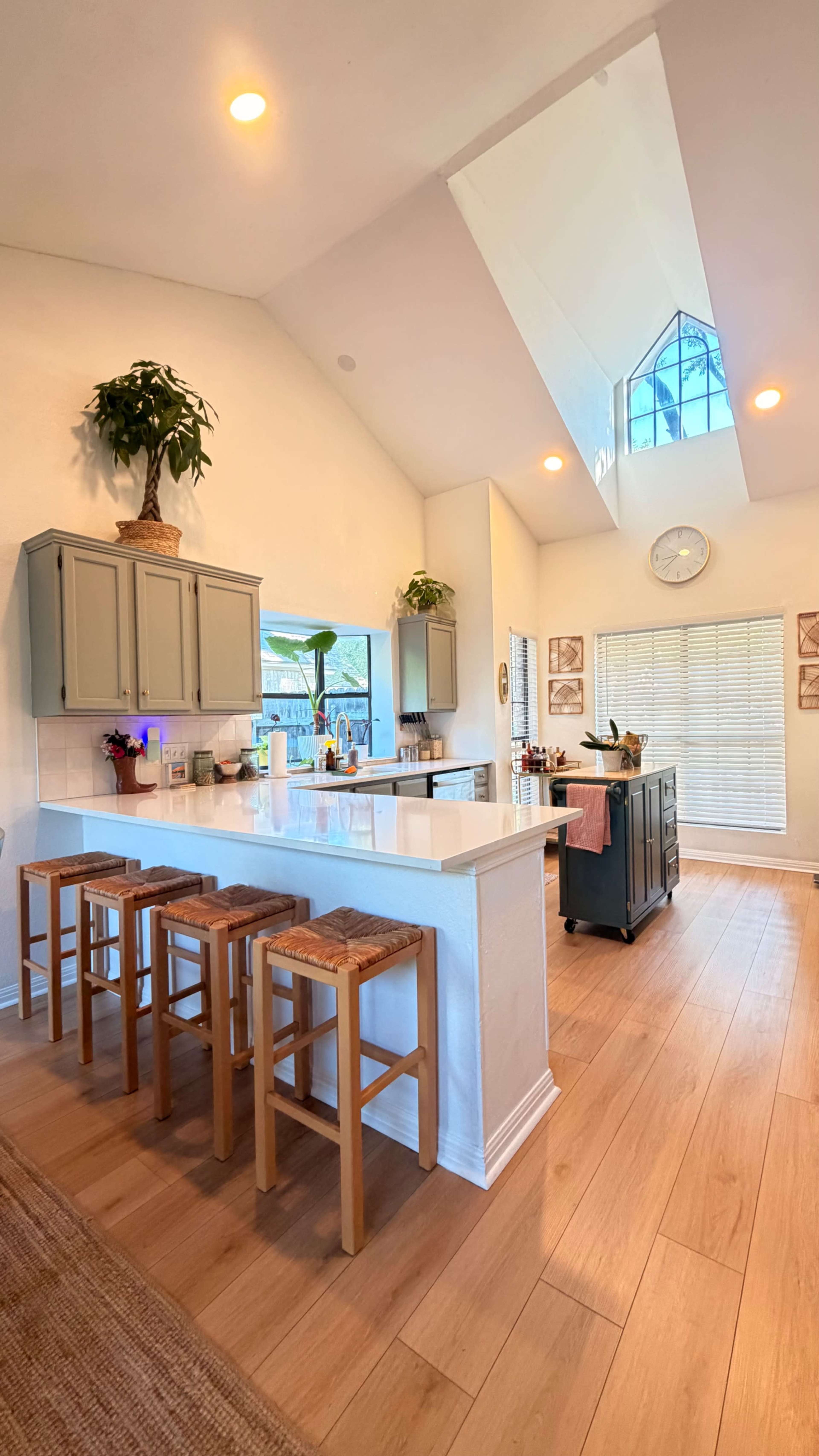 The image shows a bright kitchen with light-colored cabinets, a large island with four wooden stools, and a skylight above.