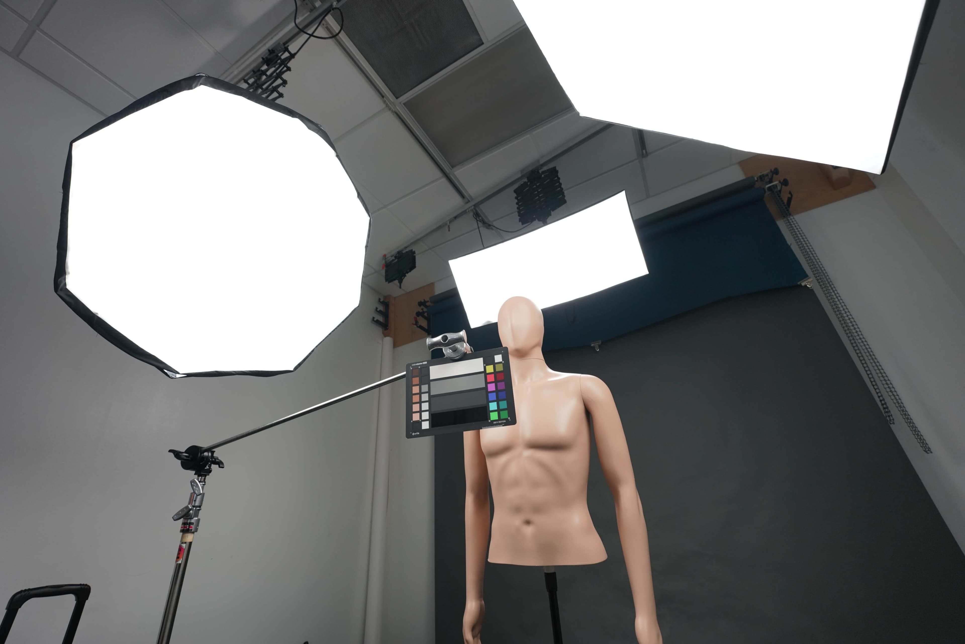 A photography studio setup features a mannequin in front of a gray backdrop, with softbox lights positioned overhead and a color reference chart attached to a camera on a tripod.