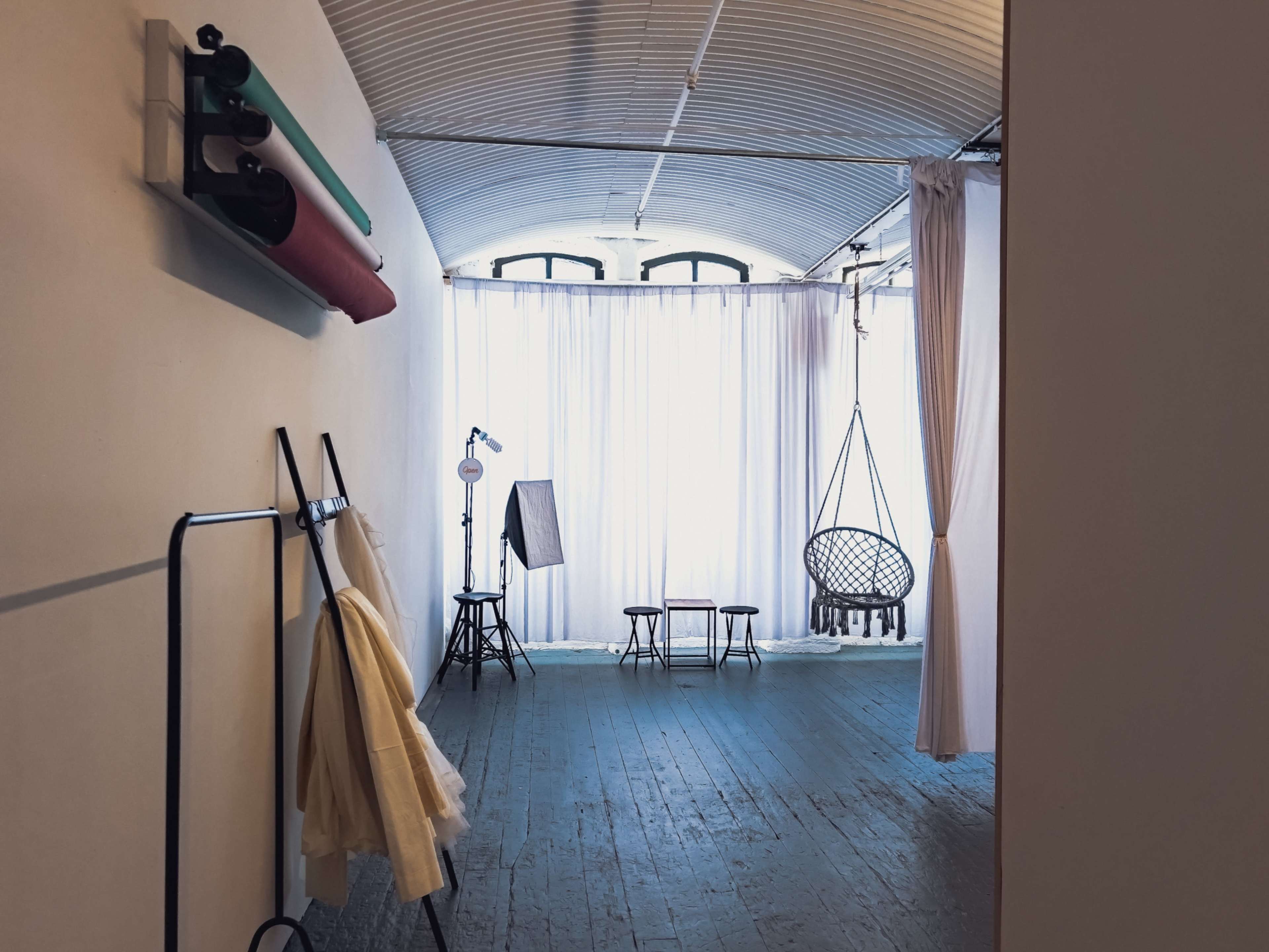 The image shows a spacious interior with large windows, featuring a hanging chair, a few stools, and a stand with fabrics against a white wall.