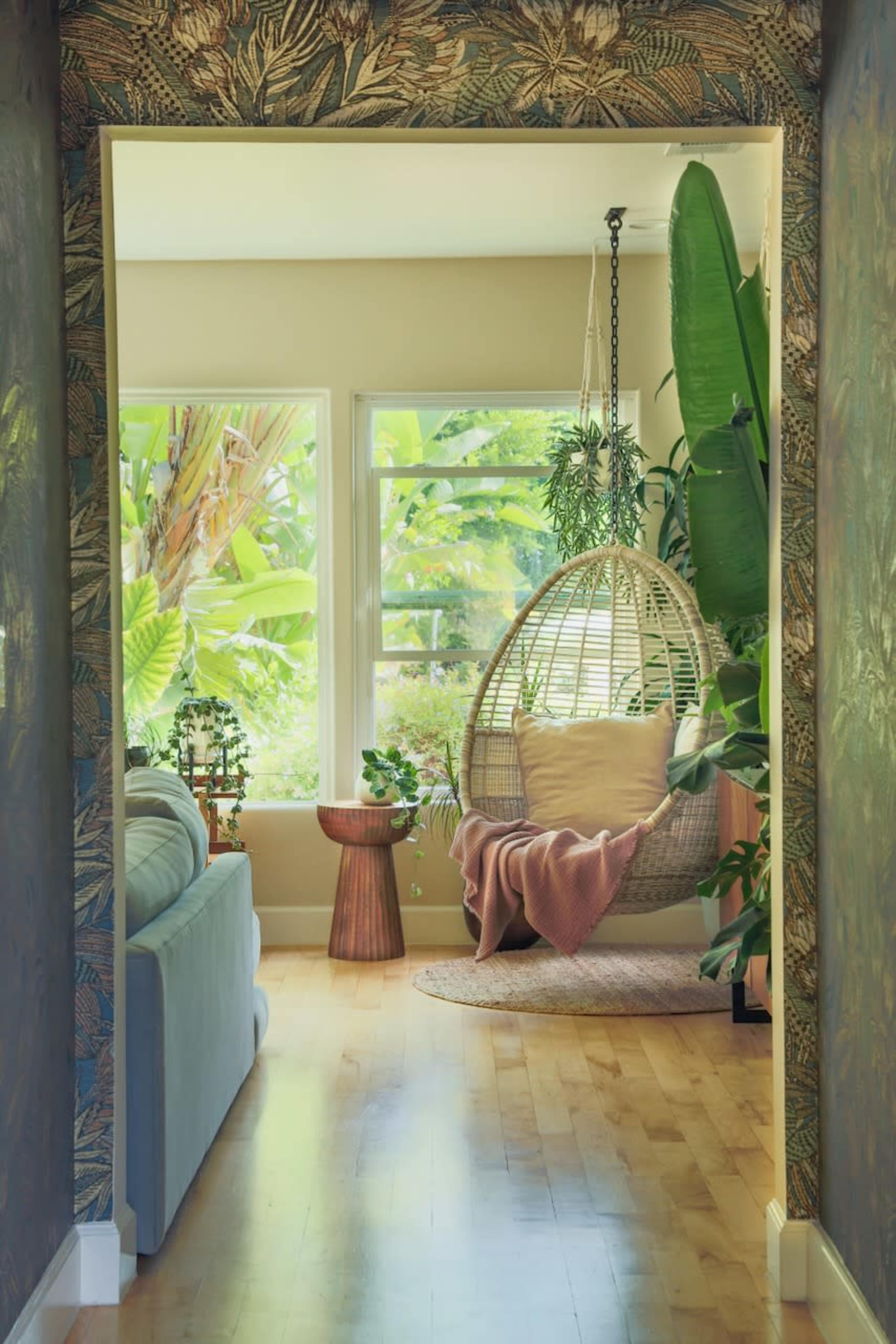A stylish indoor space features a hanging rattan chair, small wooden table, and lush greenery visible through large windows.