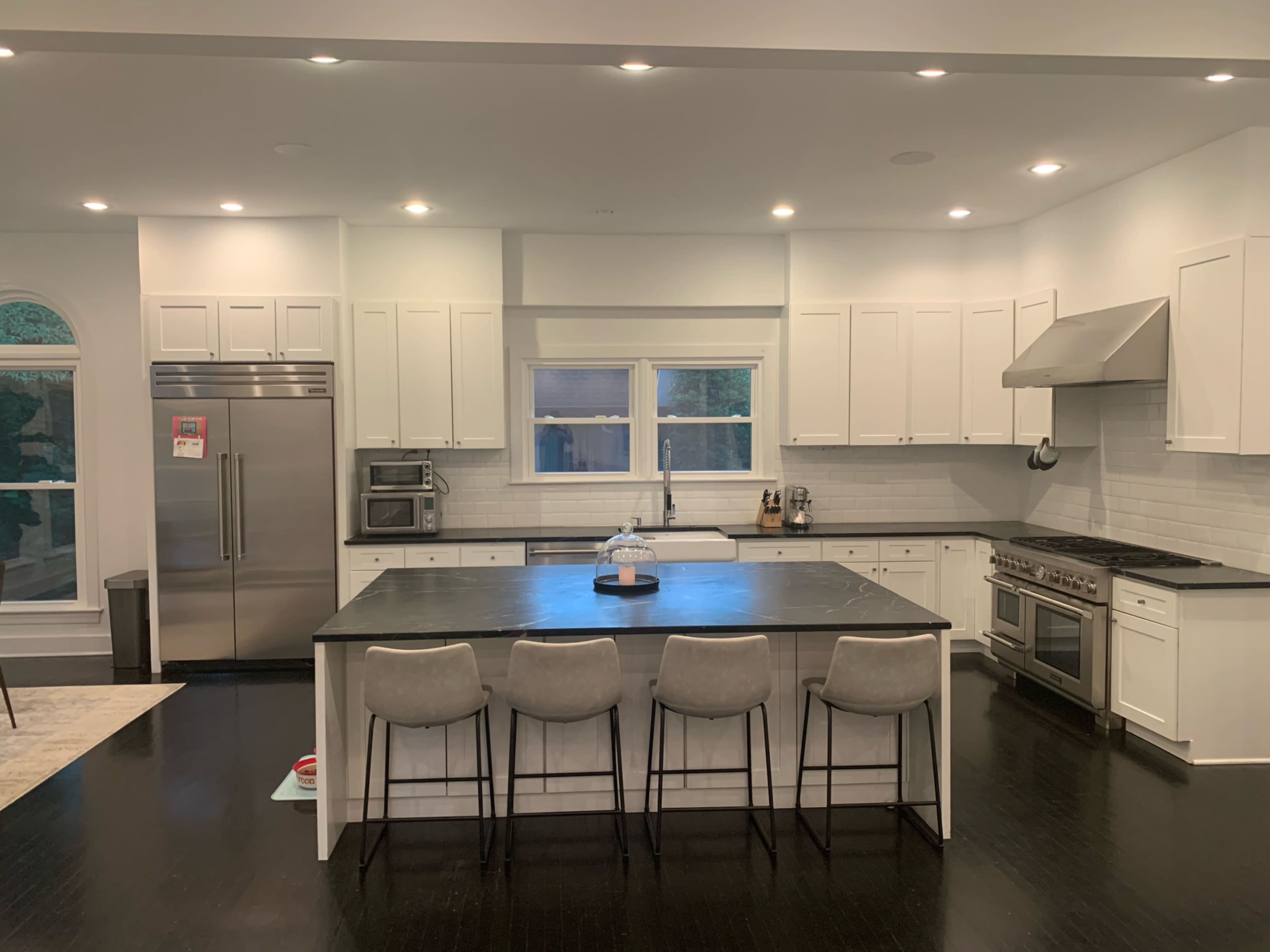 A modern kitchen features white cabinetry, a large island with seating, stainless steel appliances, and dark wood flooring.