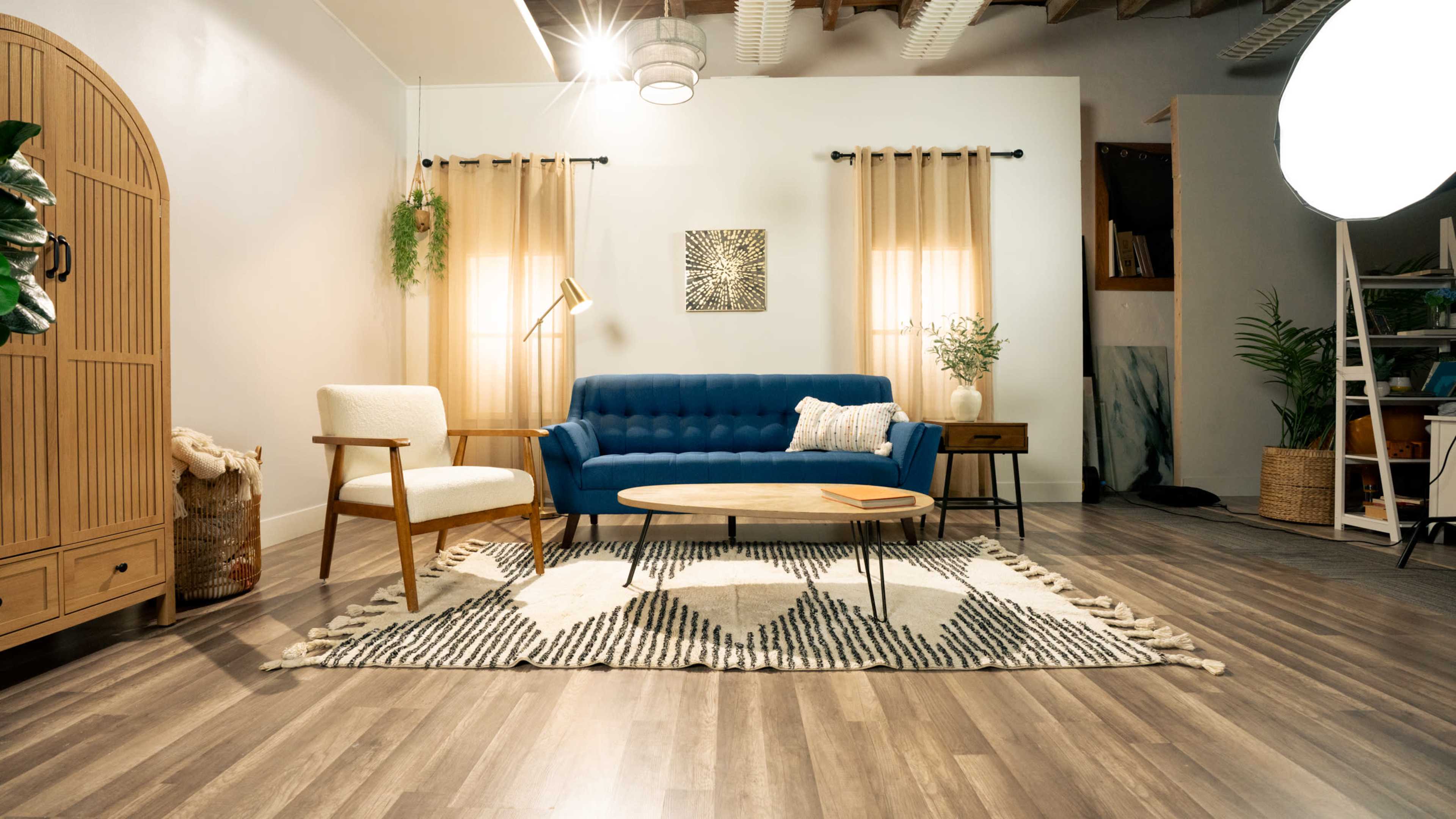 The image depicts a cozy living room interior featuring a blue sofa, an armchair, a round coffee table, and decorative plants, all arranged on a geometric patterned rug.