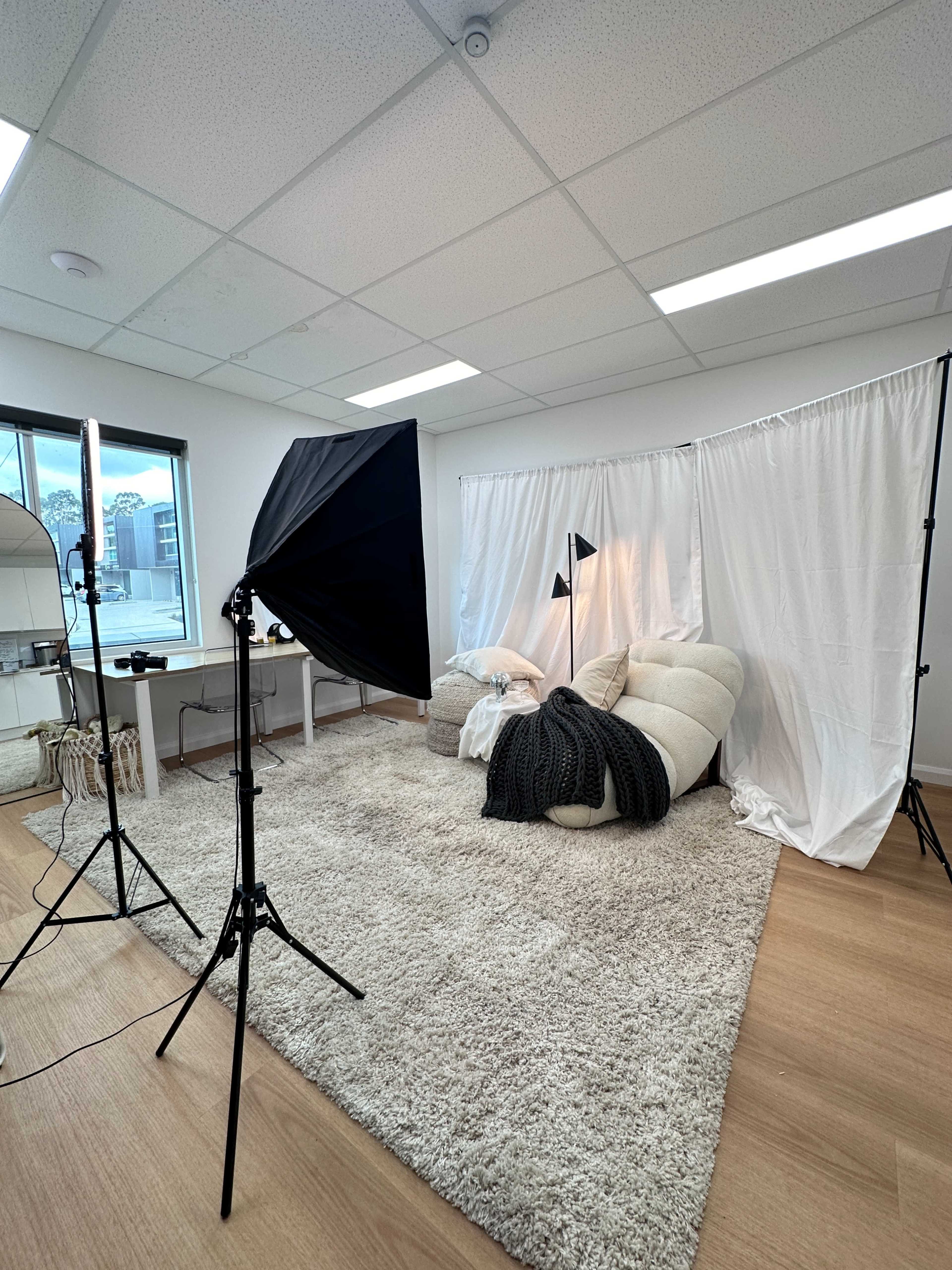 The image shows a well-lit photography studio featuring a plush chair, a decorative rug, a backdrop of white fabric, and two softbox lights set up for a photoshoot.