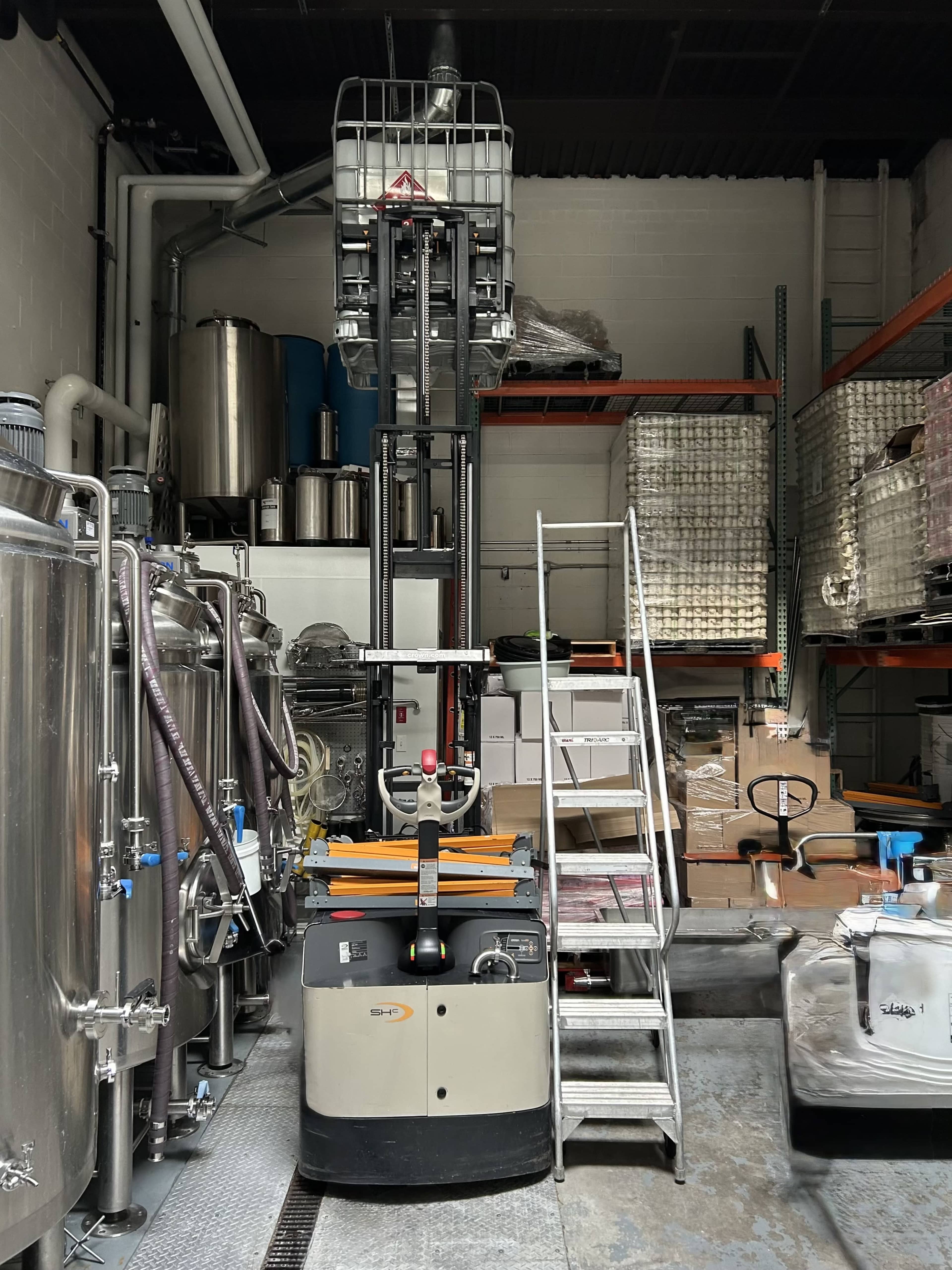 An electric forklift is positioned in a storage area, surrounded by large metal tanks and stacked shelves with various materials.