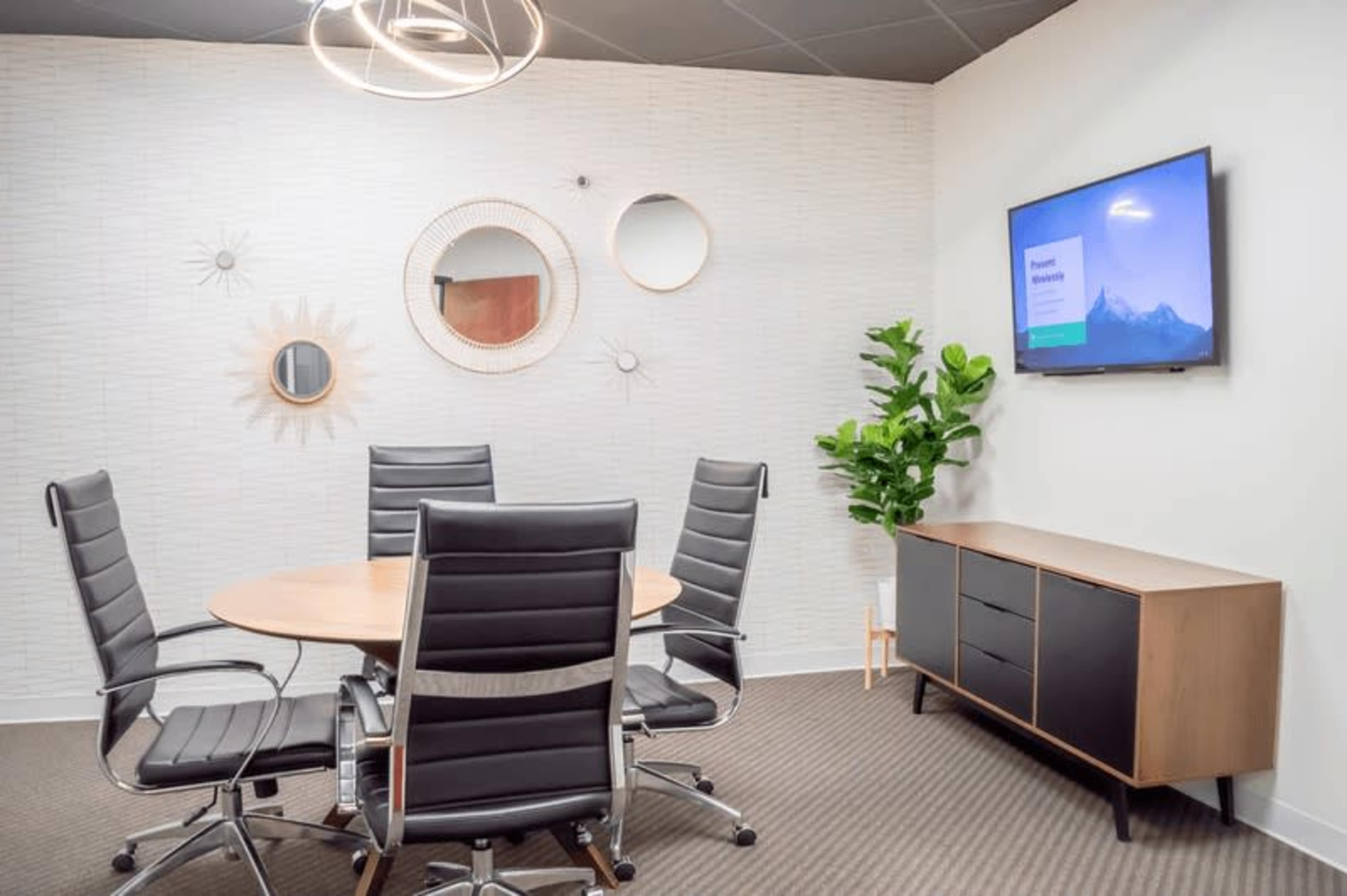 A small conference room with a round table surrounded by six black chairs, a plant, a wall-mounted TV, and decorative mirrors on the walls.