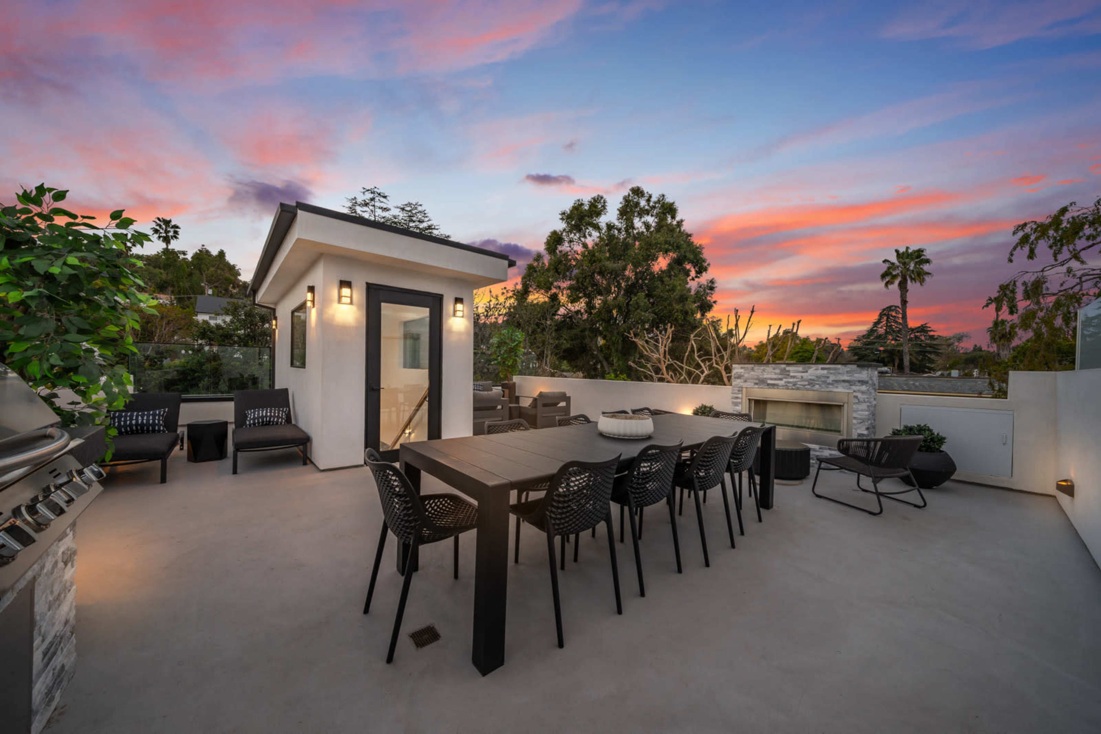 A modern outdoor dining area features a long table with black chairs, surrounded by a sleek grill and a cozy lounge space, set against a vibrant sunset sky.