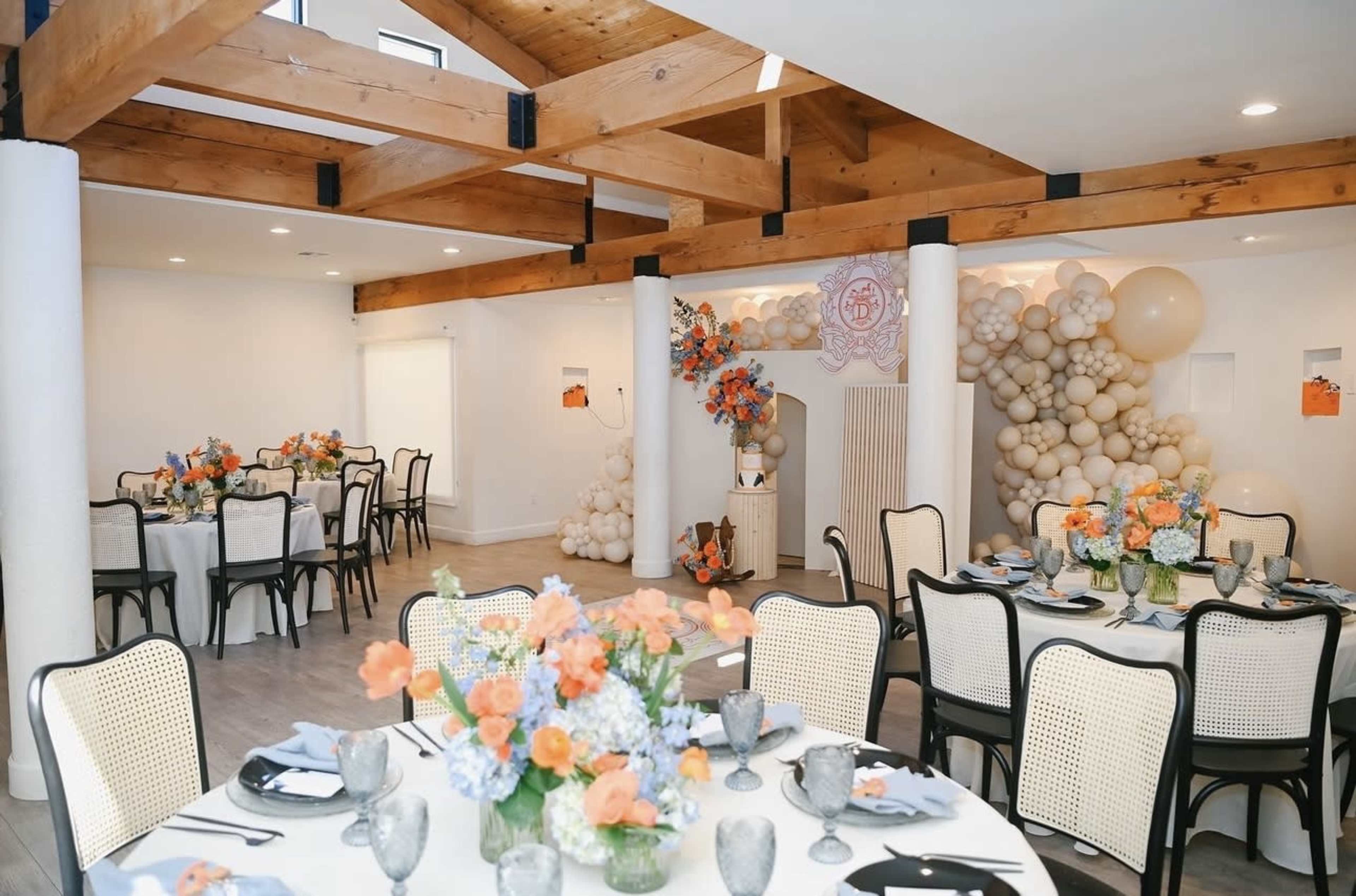 A decorated event space features tables set with floral arrangements and balloons, with wooden beams visible in the ceiling.