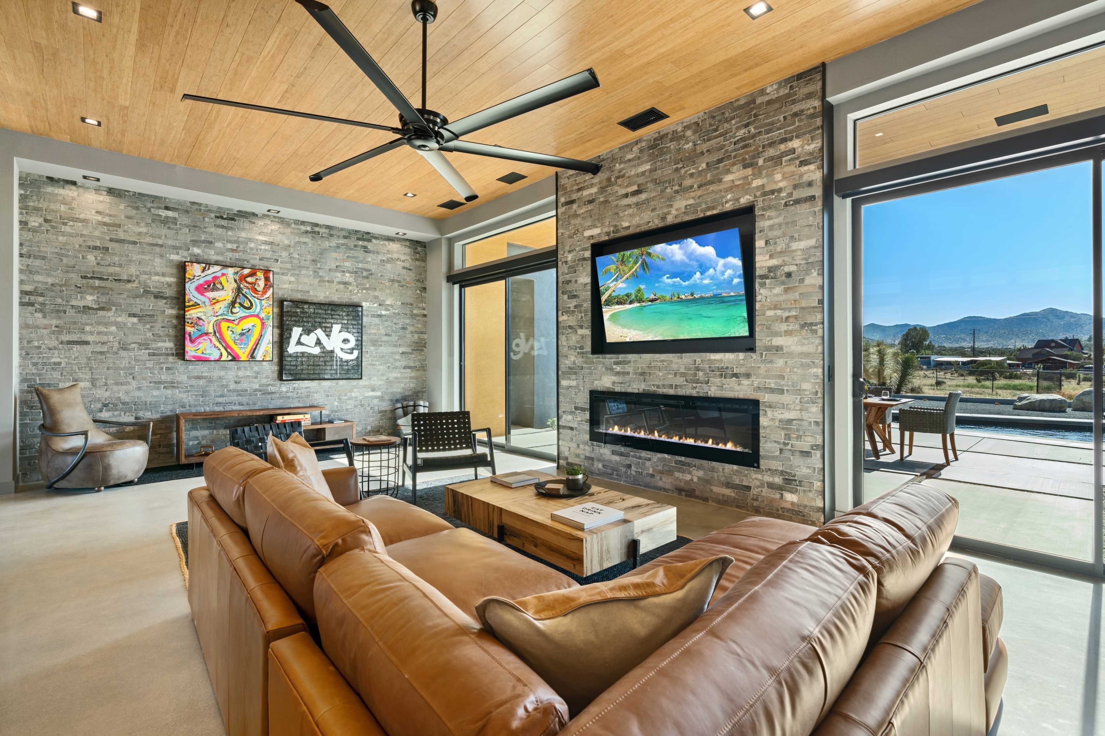 The image shows a modern living room with a leather sofa, stone accent walls, a wall-mounted TV, and large windows offering a view of the outdoor landscape.