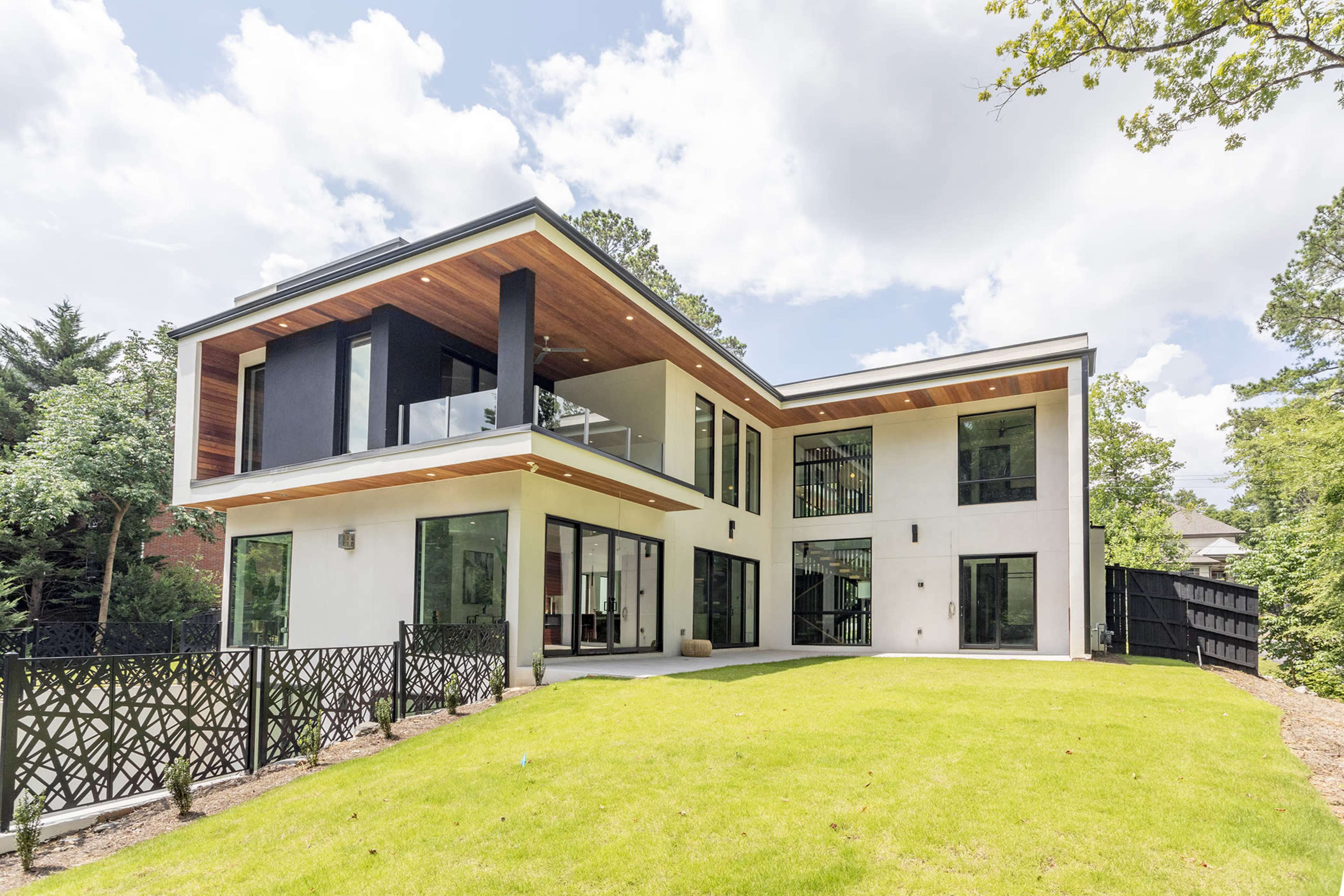 A modern two-story house with large windows and a flat roof is surrounded by a well-kept lawn and bordered by a wooden fence.