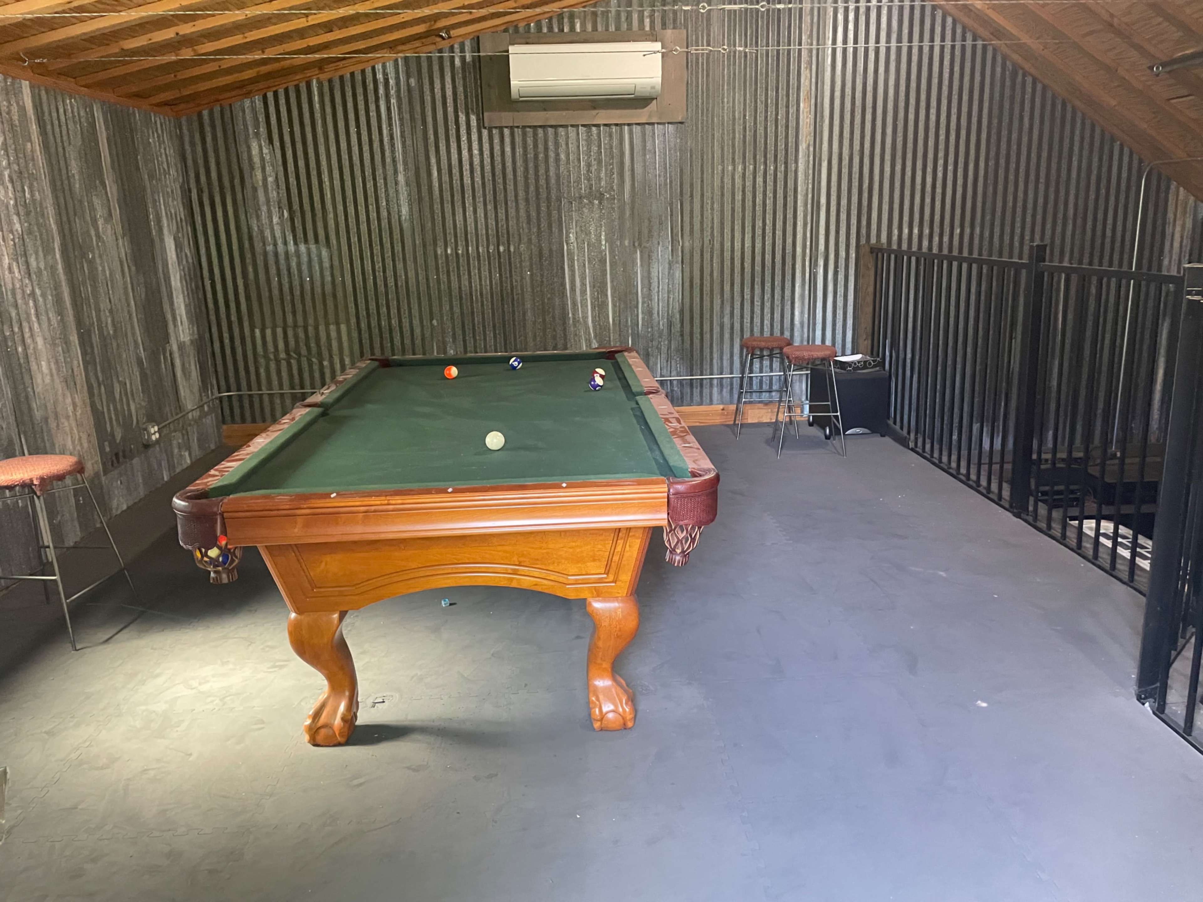 A wooden pool table sits in a room with corrugated metal walls, accompanied by several bar stools.