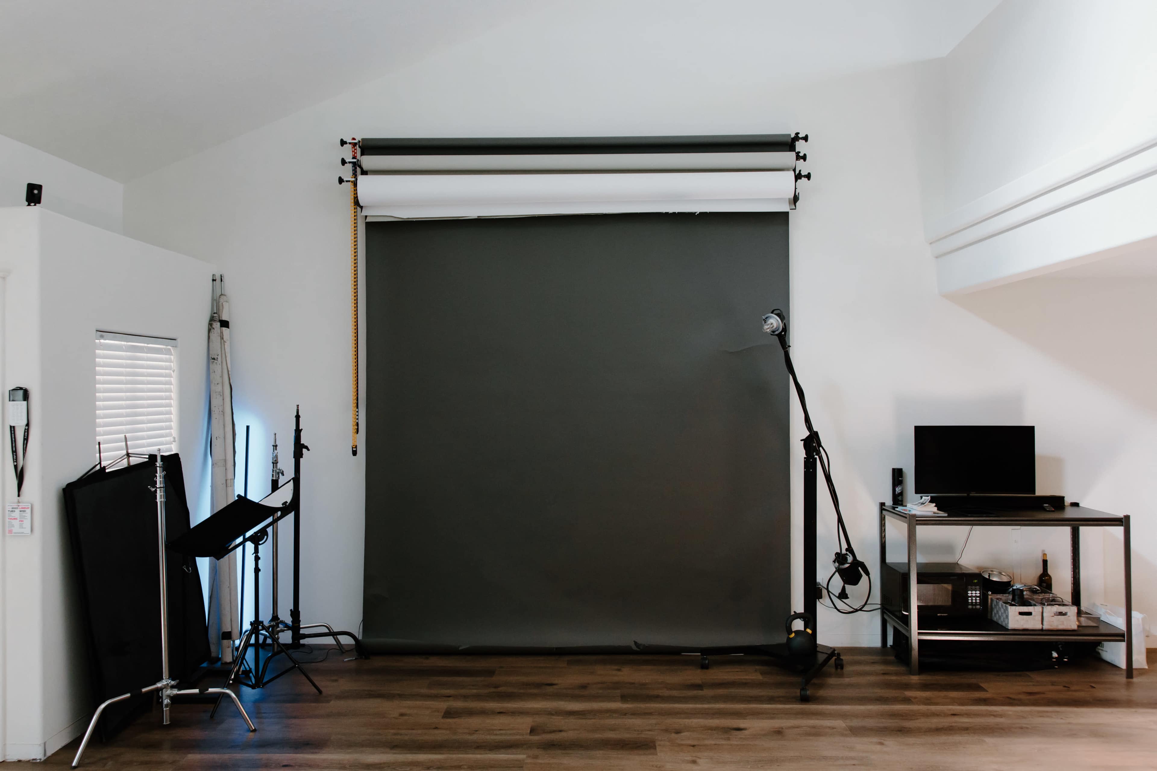 A photography studio features a green backdrop, lighting equipment, and a monitor on a stand against a white wall.
