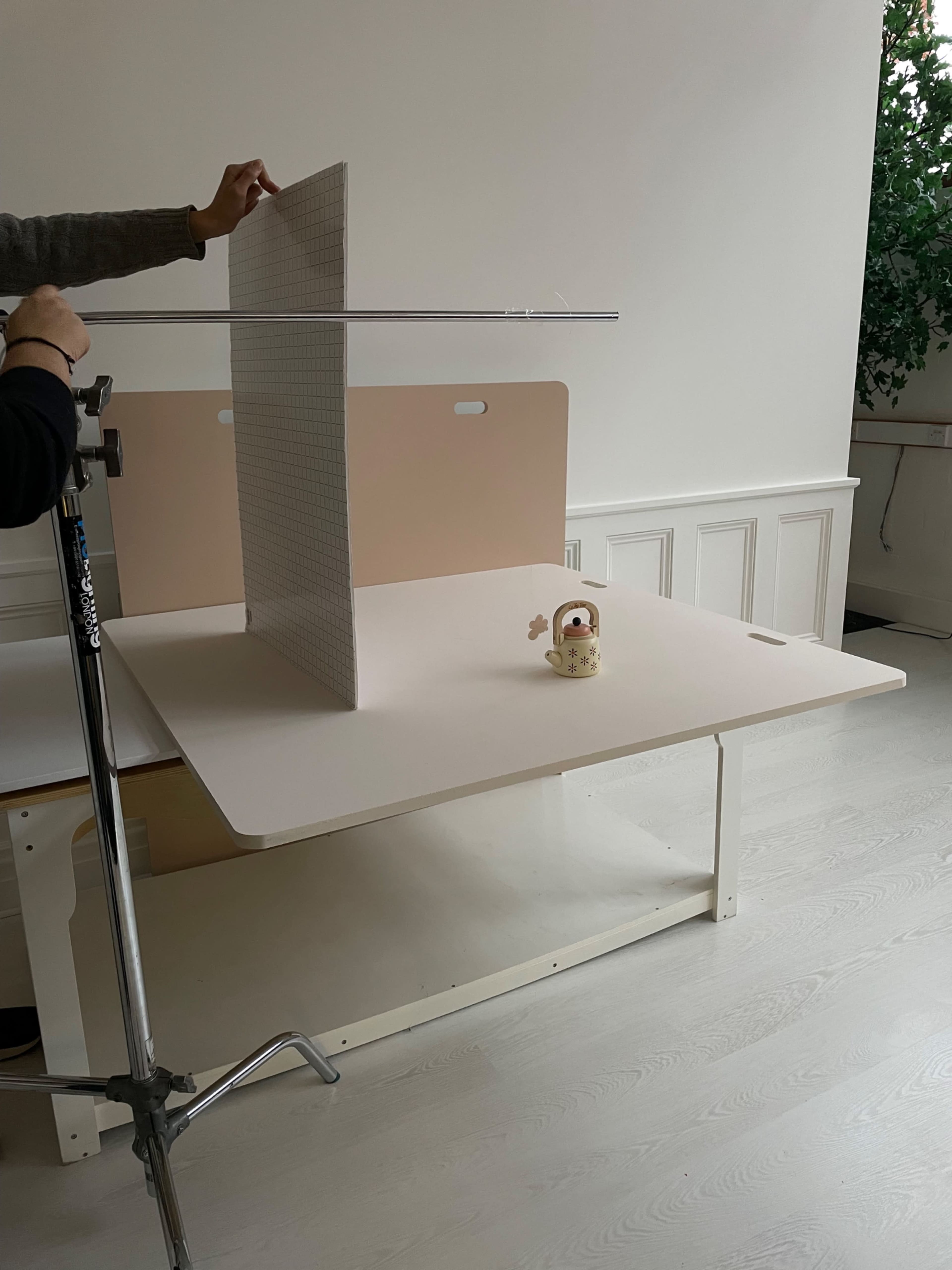 A person adjusts a light stand with a reflector near a small decorative object on a table in a bright room.