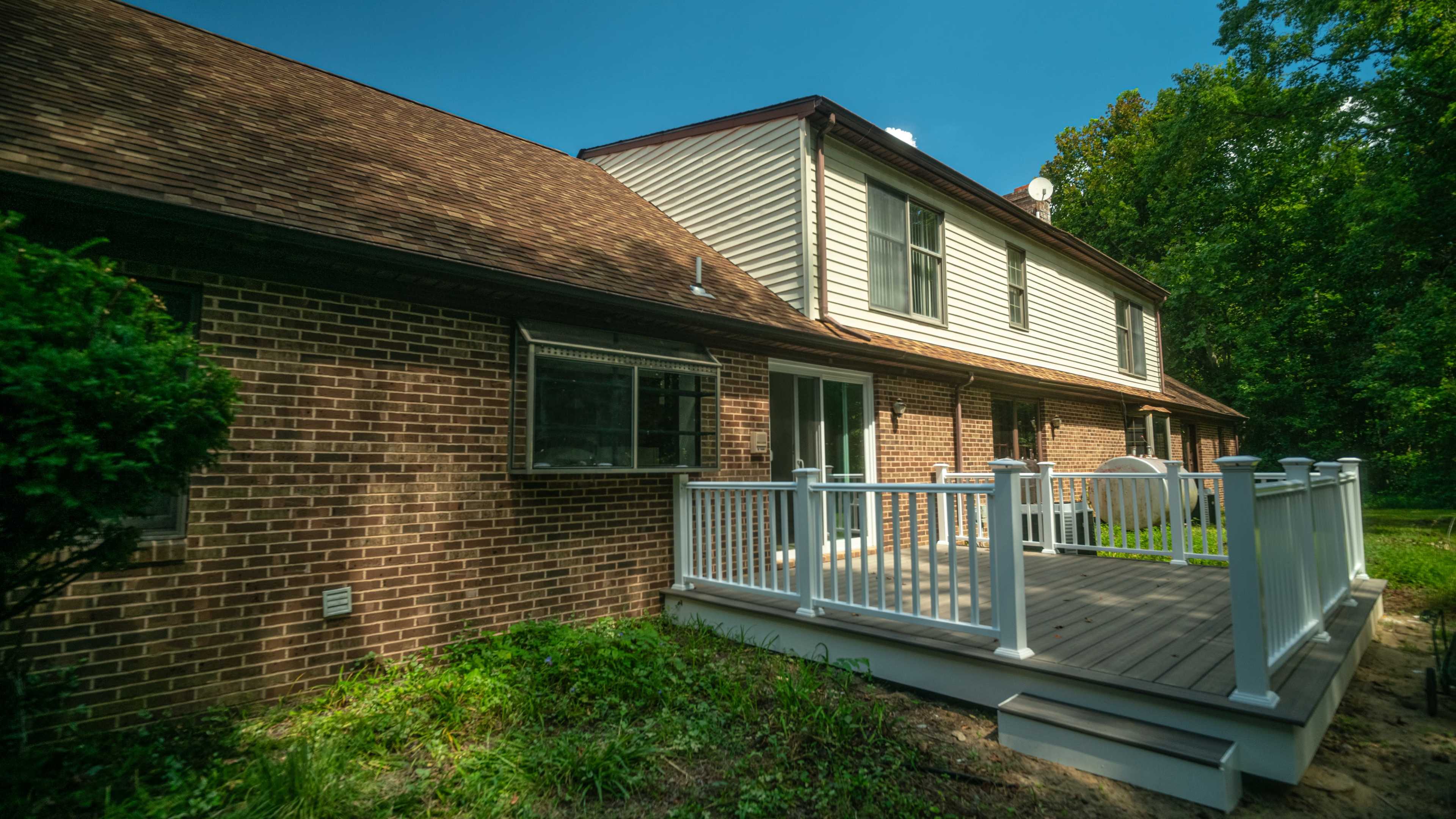The image shows the exterior of a two-story brick house with a wooden deck and railing, surrounded by greenery.