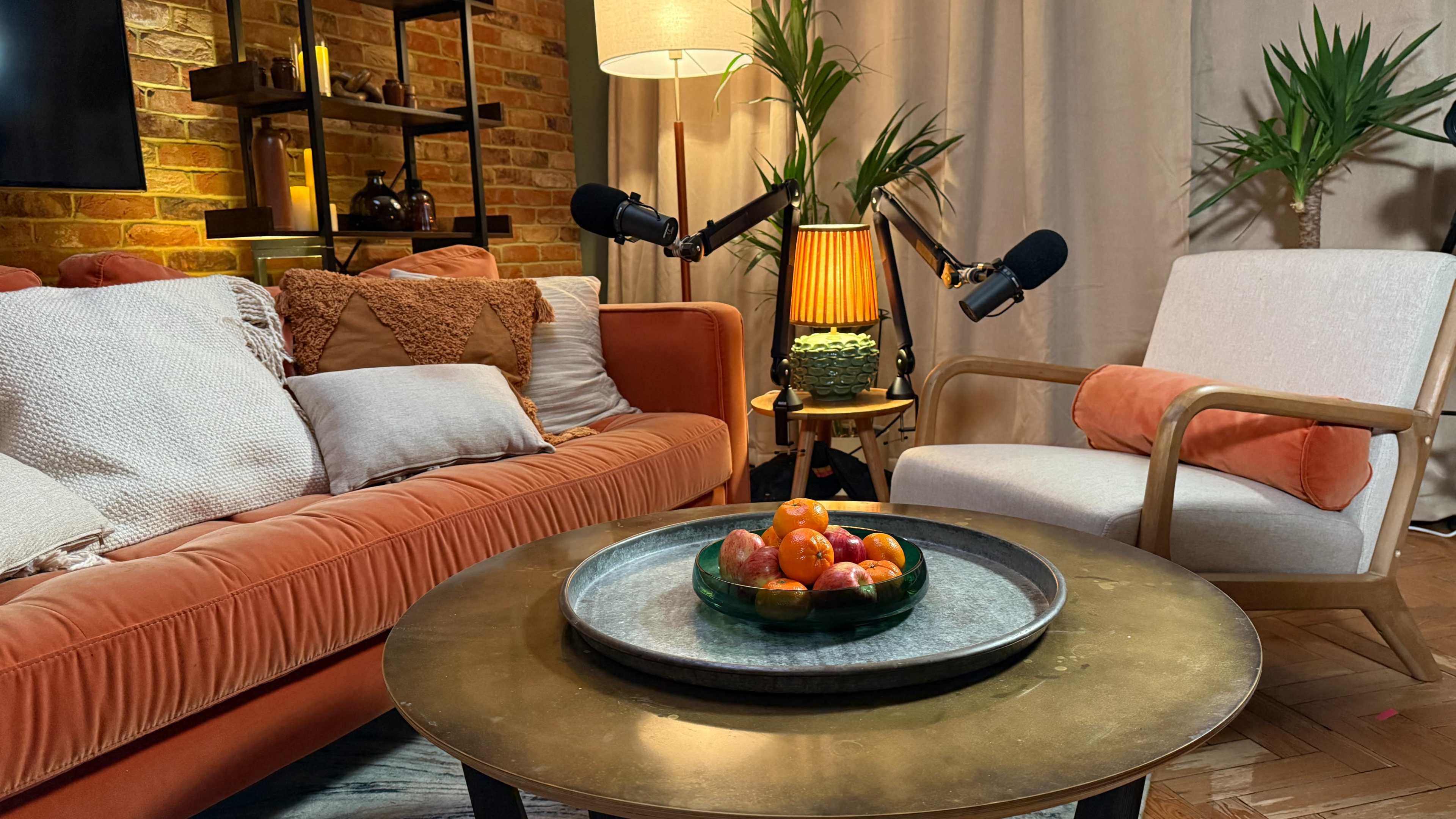 A cozy living room scene features an orange sofa, a round coffee table with a bowl of fruit, and a chair beside a potted plant, all illuminated by warm lighting.