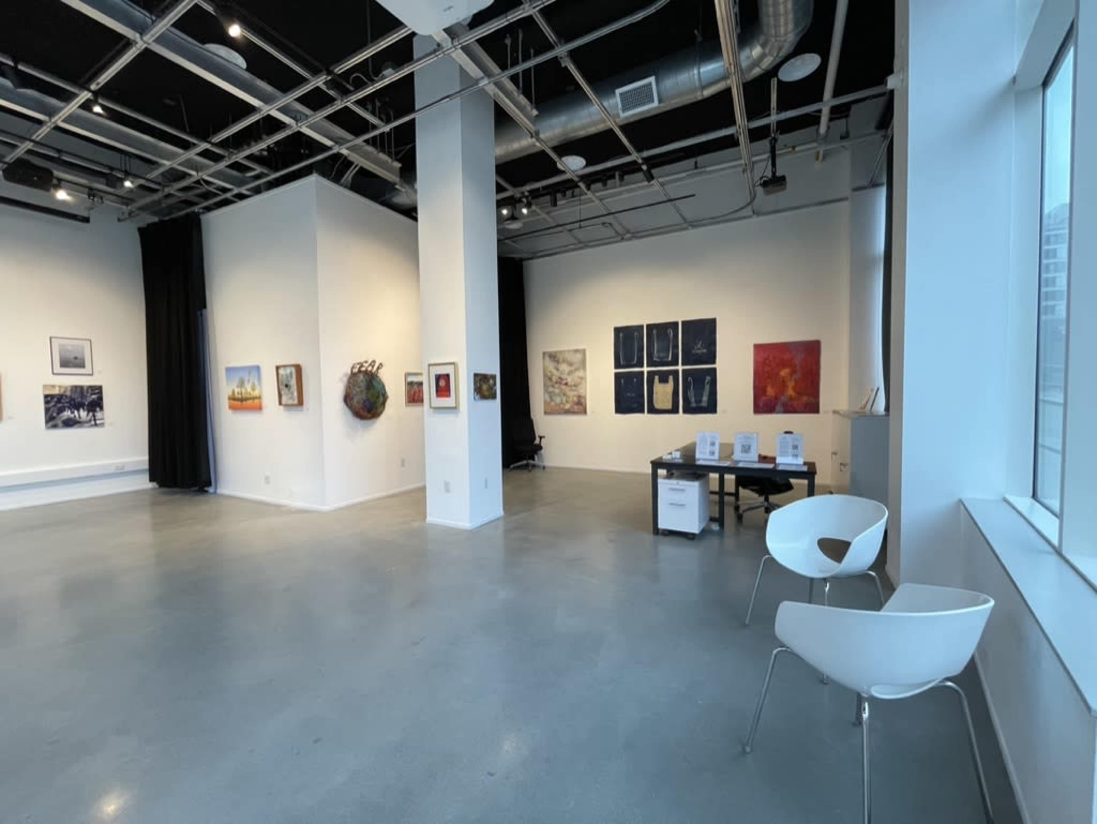 The image shows a spacious art gallery with various artworks displayed on white walls, a desk in the corner, and a chair near a window.