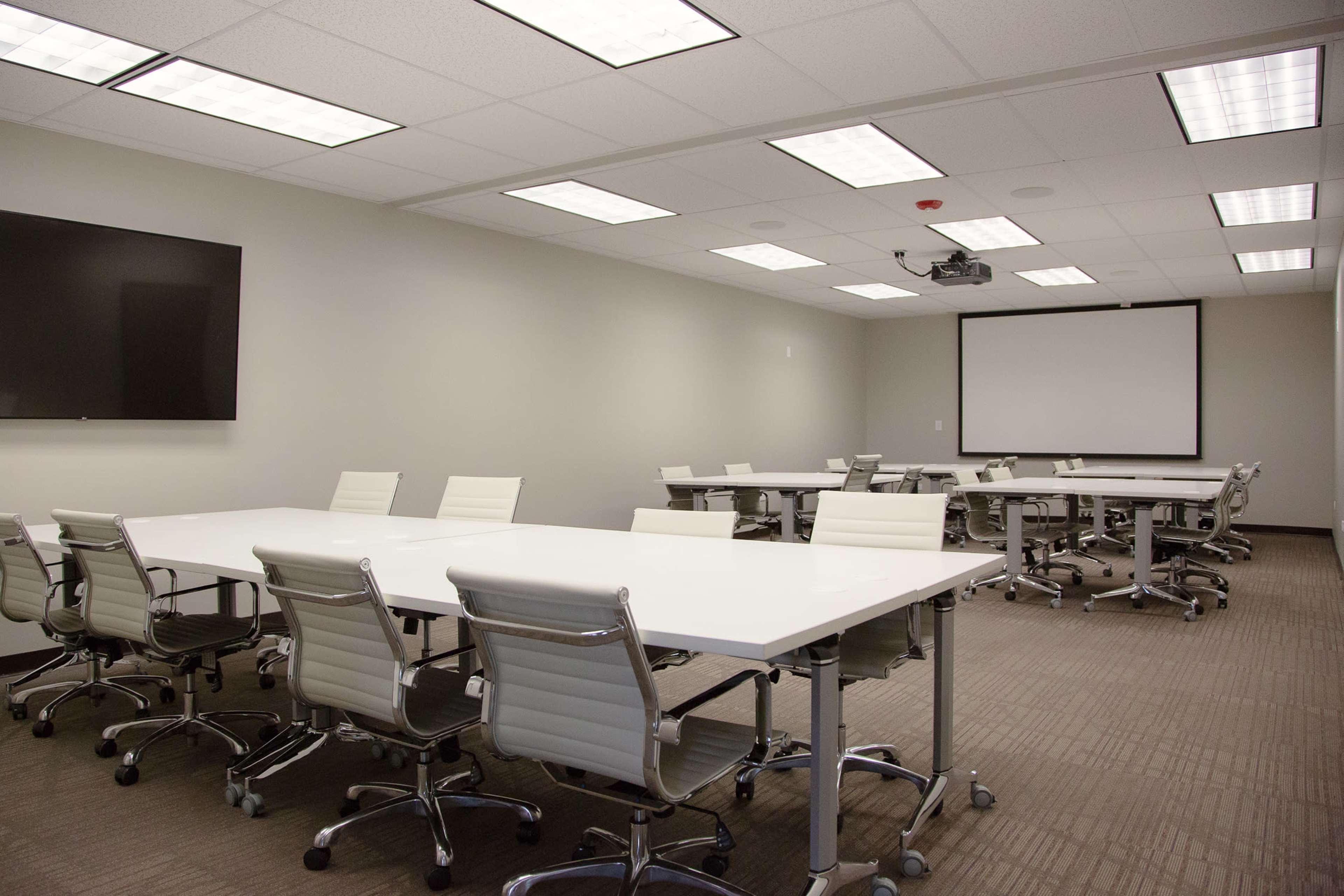 A modern meeting room with multiple white tables and chairs arranged in a symmetrical layout, equipped with a projector and two screens mounted on the walls.