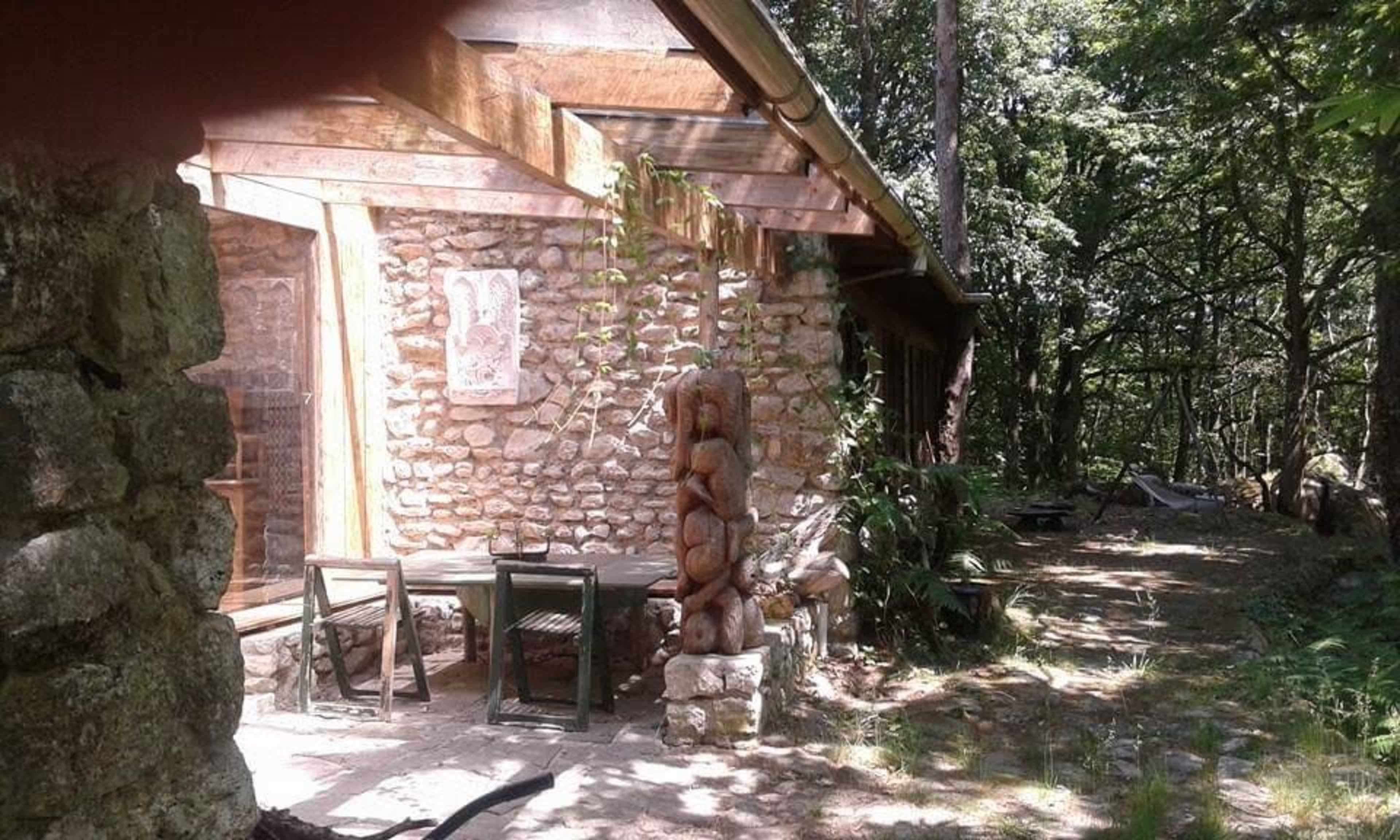 A stone and wood cabin is surrounded by trees, featuring a carved wooden sculpture and a table set on the patio.