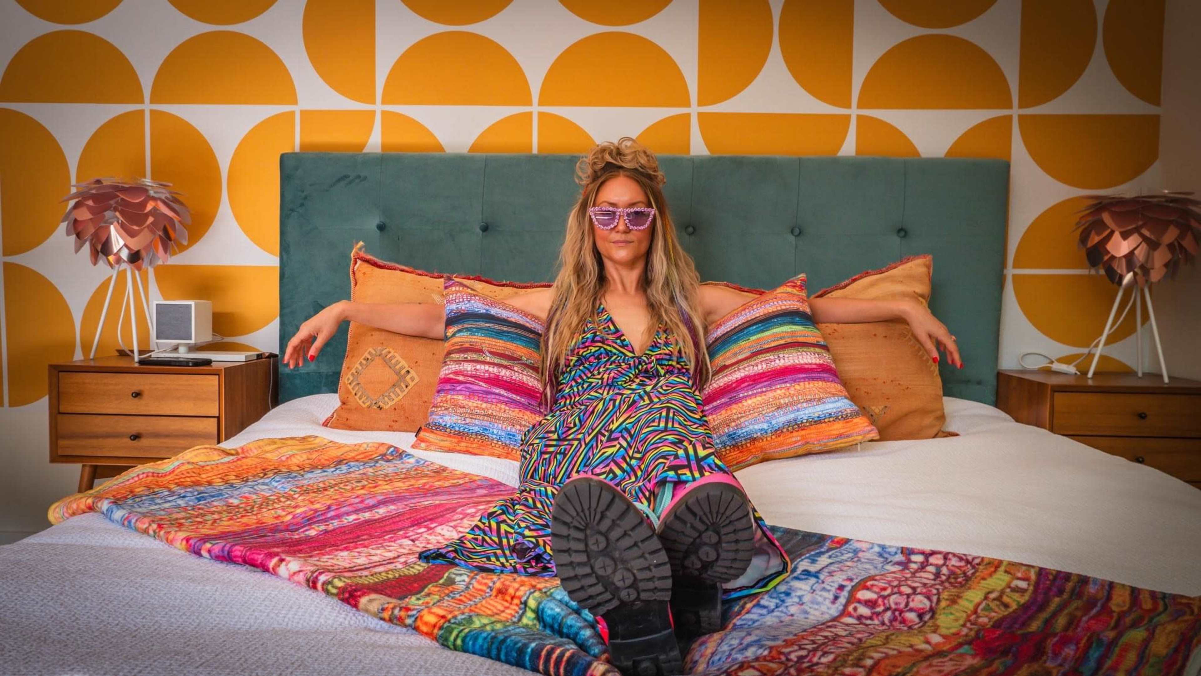 A woman in a colorful dress sits on a bed with vibrant patterned pillows and a textured throw, posing against a backdrop of circular wall designs.