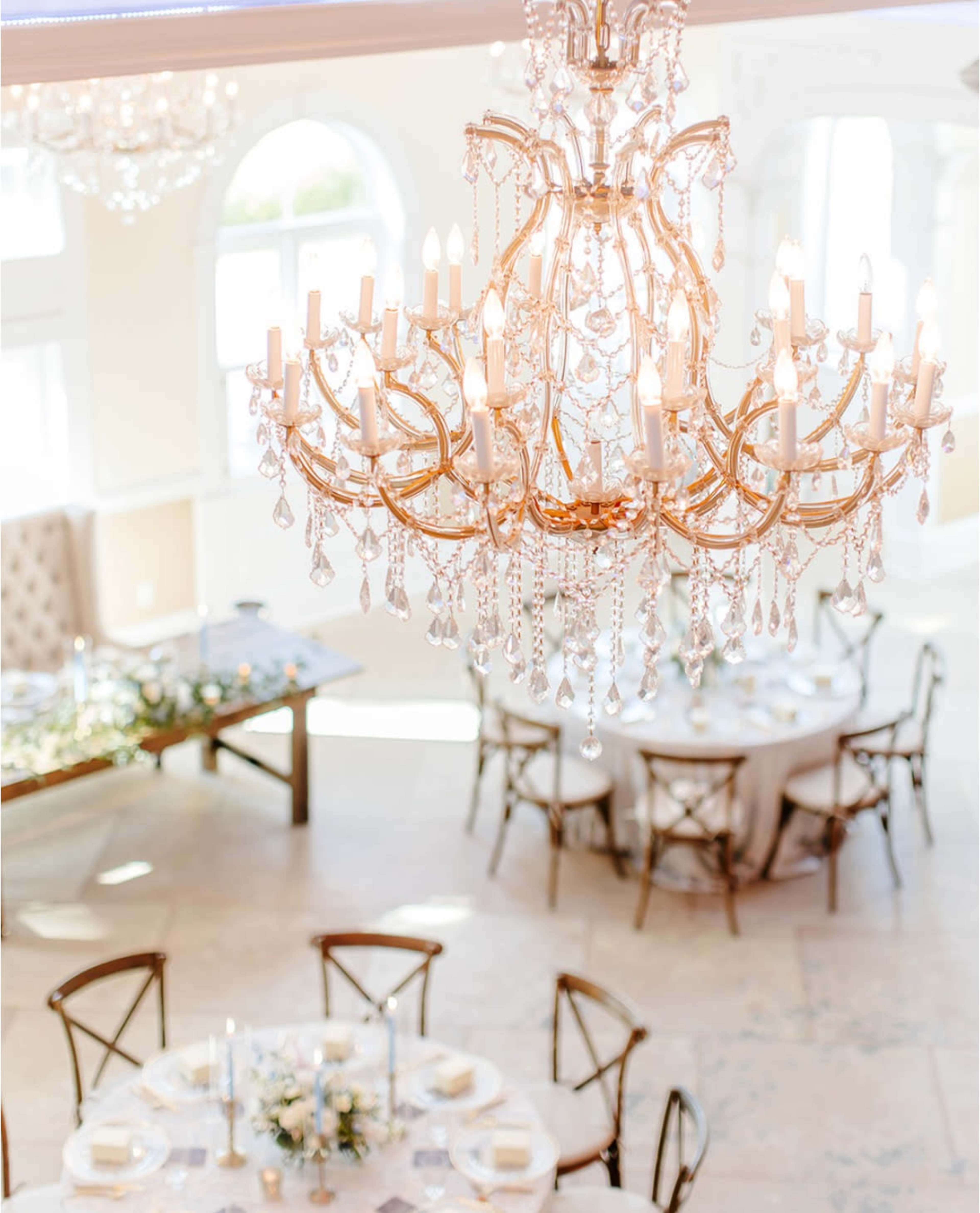 A crystal chandelier hangs from the ceiling above elegantly set tables in a well-lit dining area.