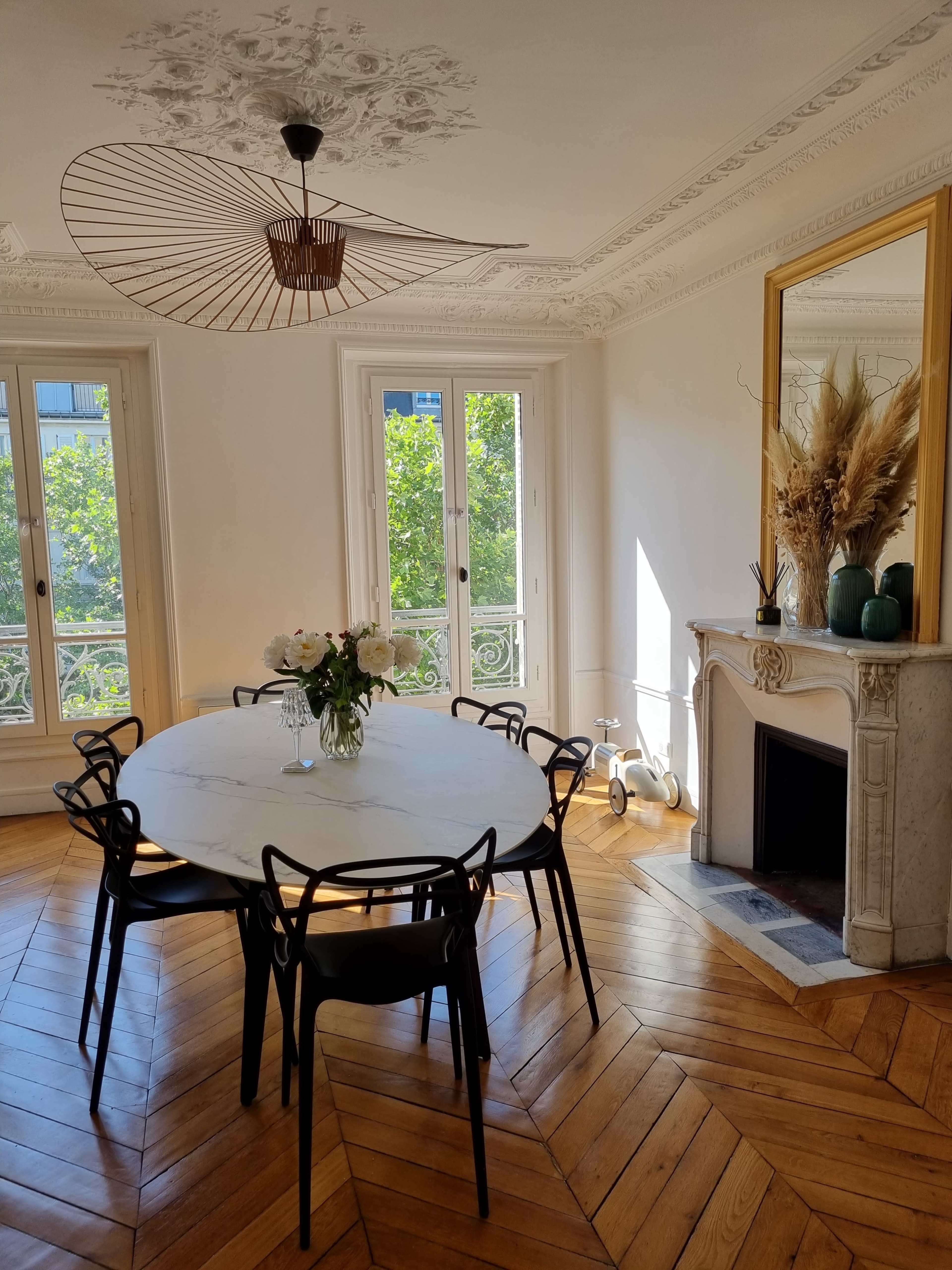A round marble dining table surrounded by black chairs is set in a bright room with large windows, a fireplace, and decorative elements.