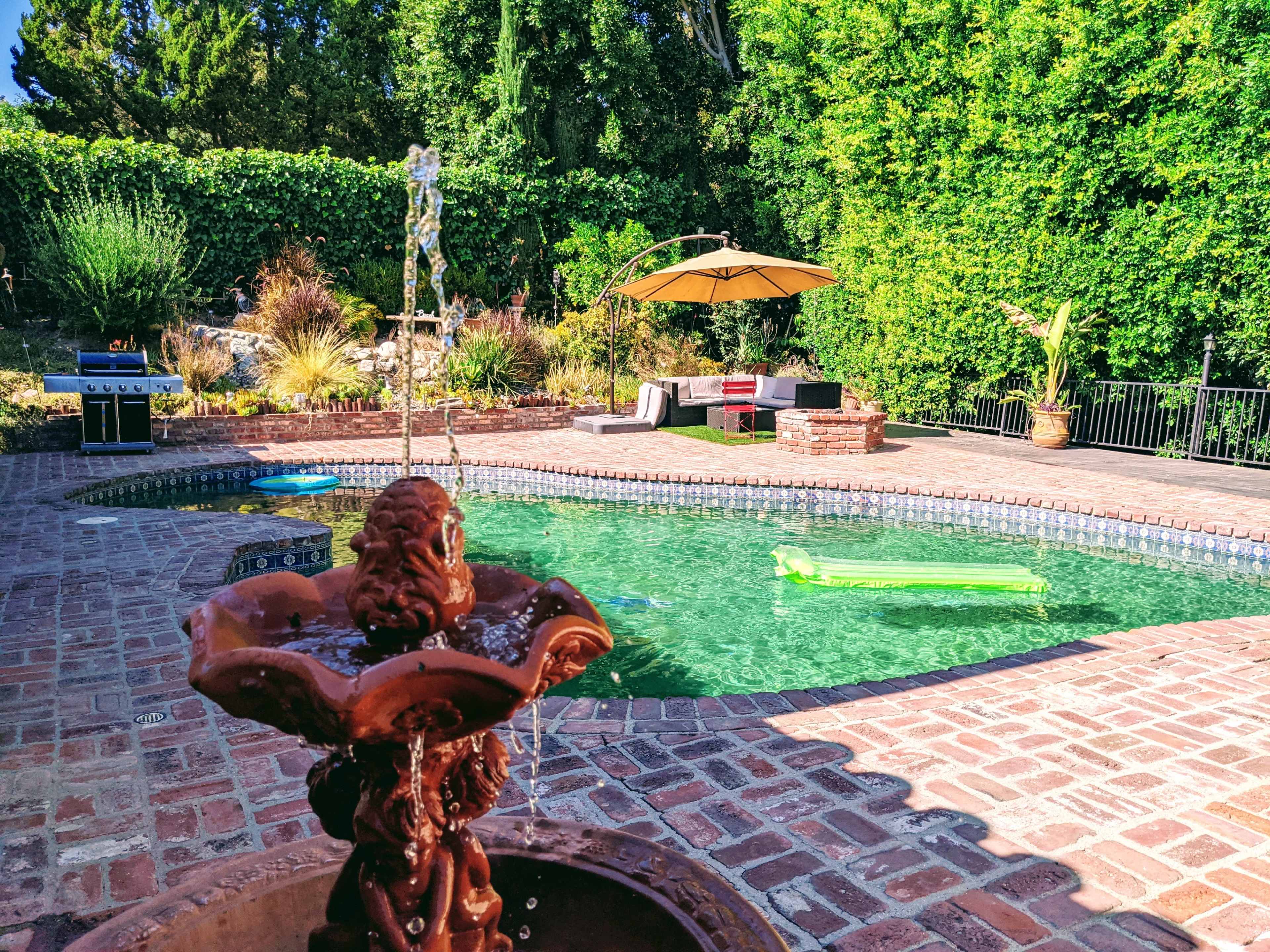 A large swimming pool with a brick patio features a fountain in the foreground, surrounded by lounge chairs and a shaded seating area.