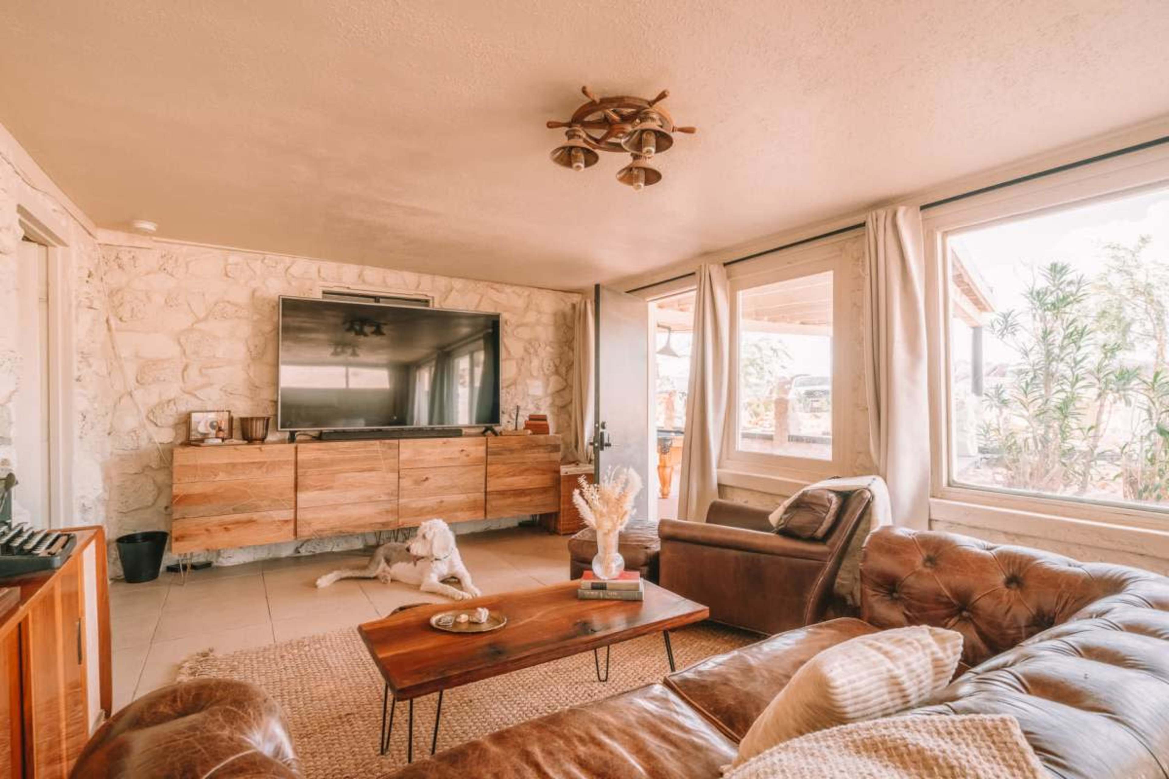 The living room features a large television mounted on a stone wall, with a wooden cabinet beneath it, a leather couch, and large windows allowing natural light to enter the space.