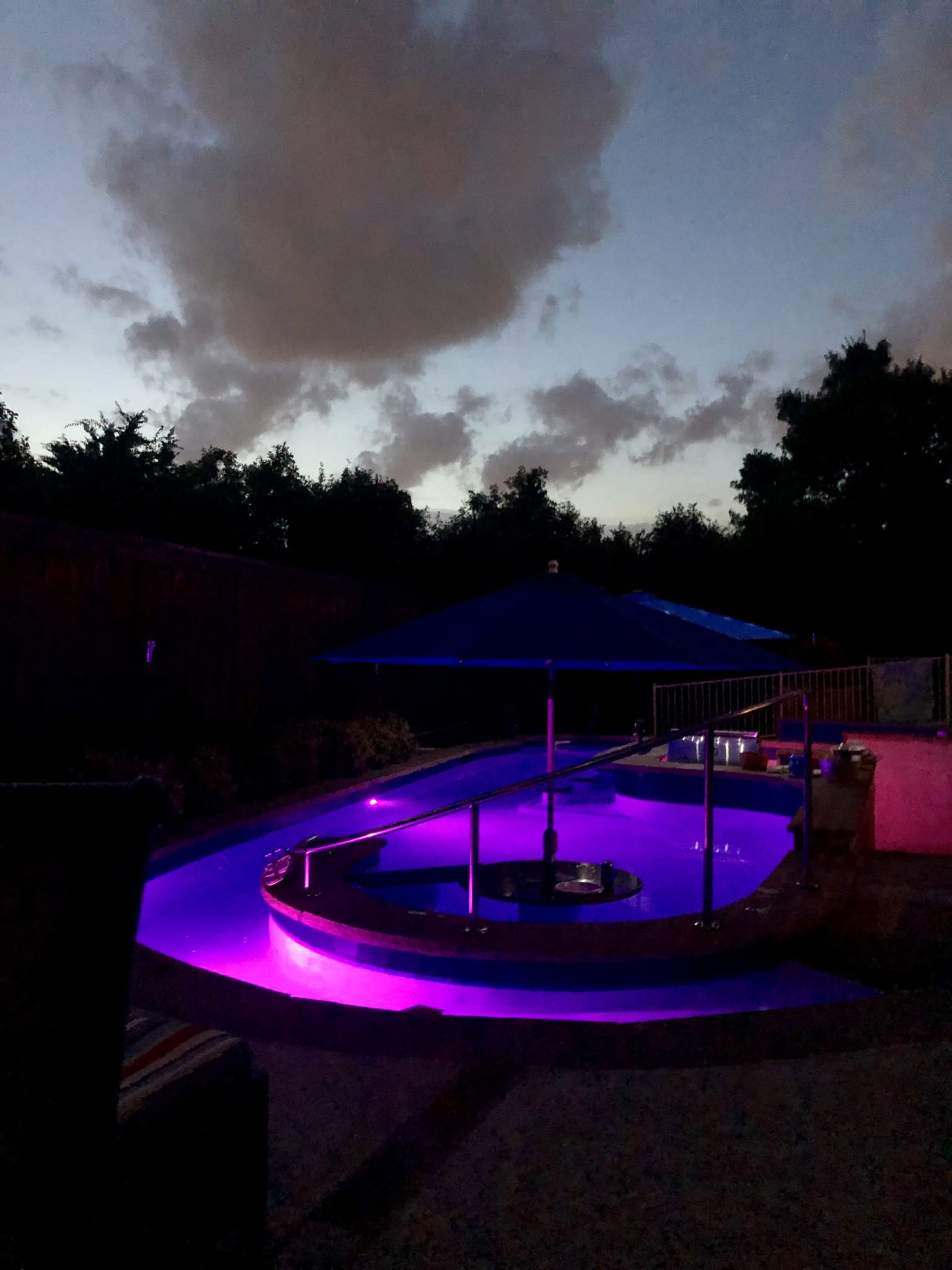 A circular pool with purple lighting is surrounded by chairs and an umbrella under a twilight sky.