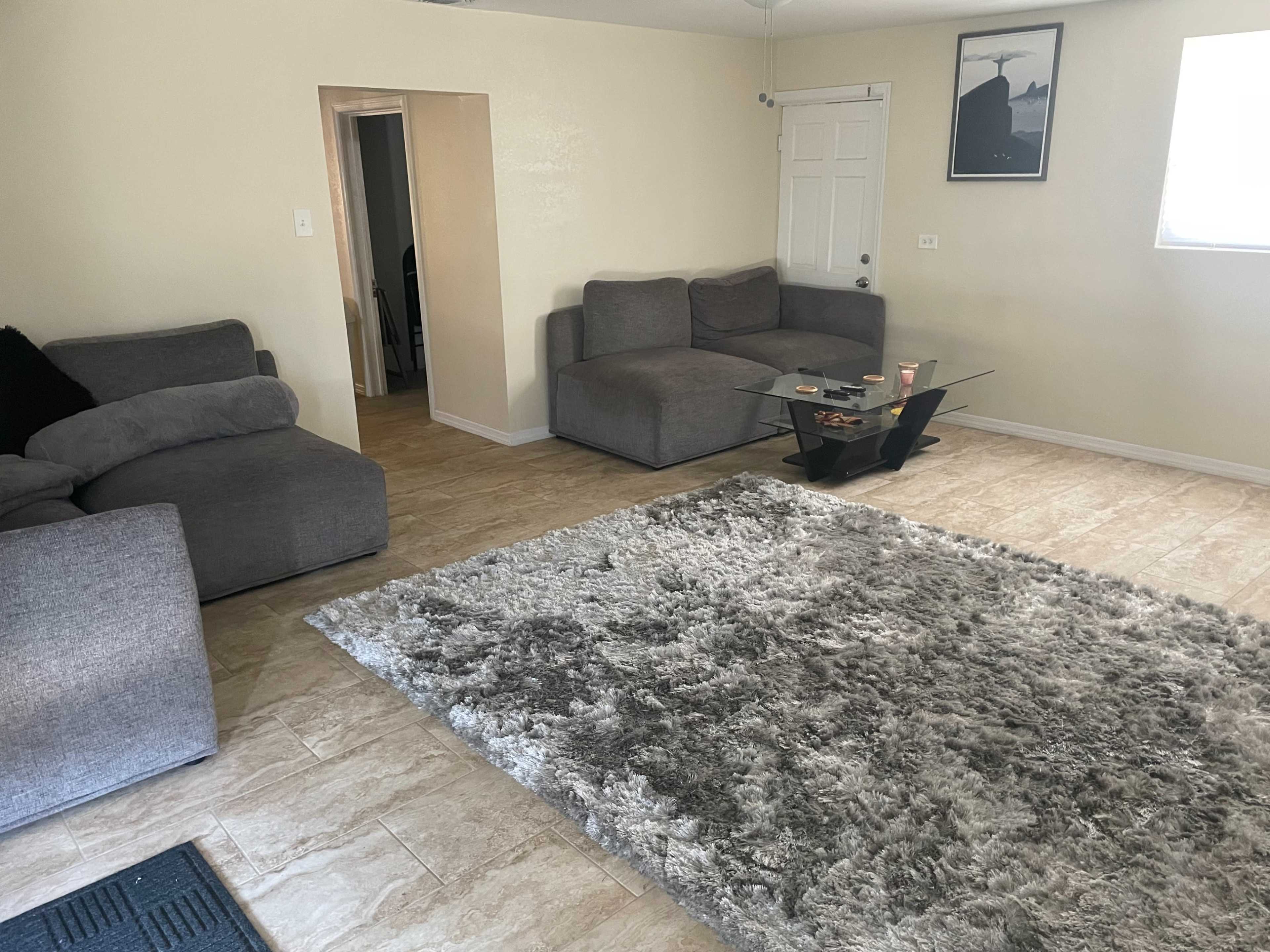 A living room with gray sofas, a glass coffee table, and a large area rug on tiled flooring.
