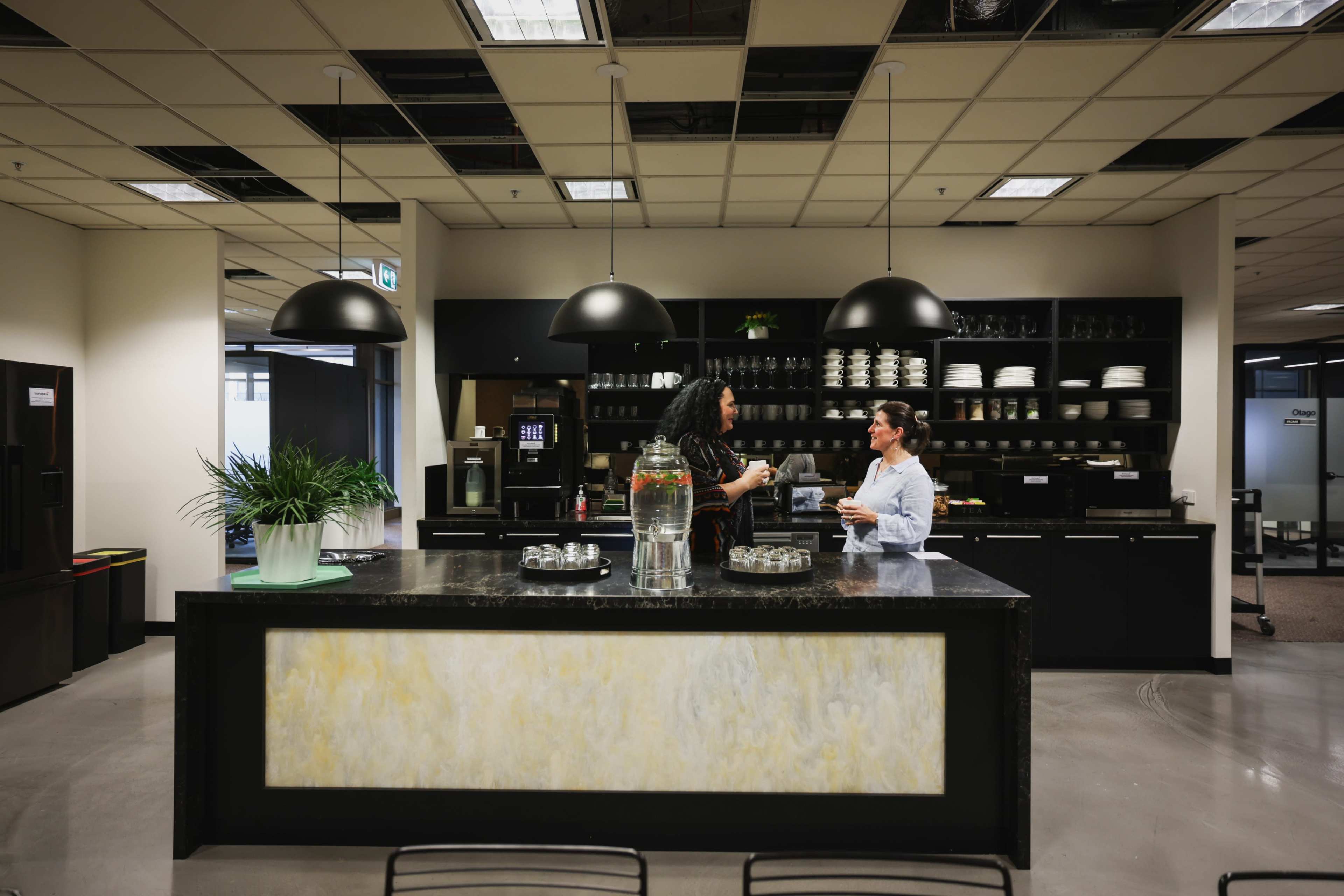Two people are engaged in conversation at a modern kitchen island in an office space, which features dark cabinetry and pendant lighting.